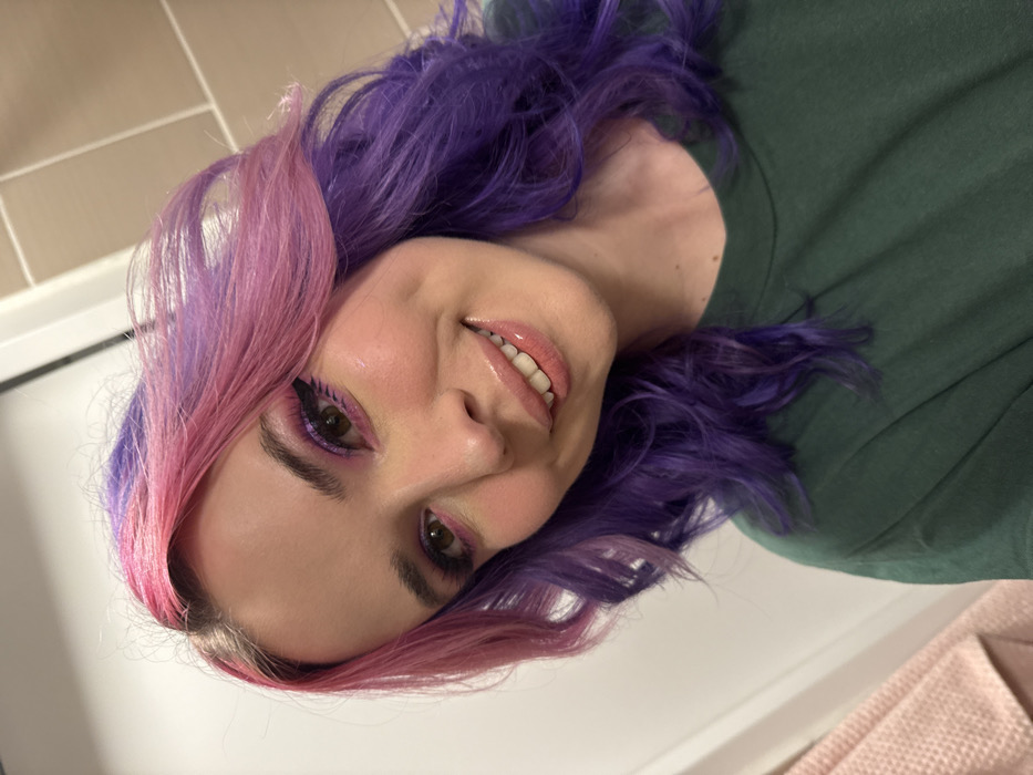 A female individual is depicted in a close-up, frontal portrait. She has wavy hair, split-dyed with pink on the viewer's left side and purple on the viewer's right side. Her eyes are made up with pink and purple eyeshadow, winged eyeliner, and dark lashes. Her lips are glossy and slightly parted in a smile, revealing her upper teeth. A dimple is visible on her right cheek. She is wearing a dark green, crew-neck t-shirt. The background consists of a plain light-colored vertical surface to the viewer's left and light brown rectangular wall tiles to the viewer's right. A pink, textured textile is partially visible in the lower-left foreground. The scene is illuminated by interior lighting. Location: City of Zagreb, Croatia.