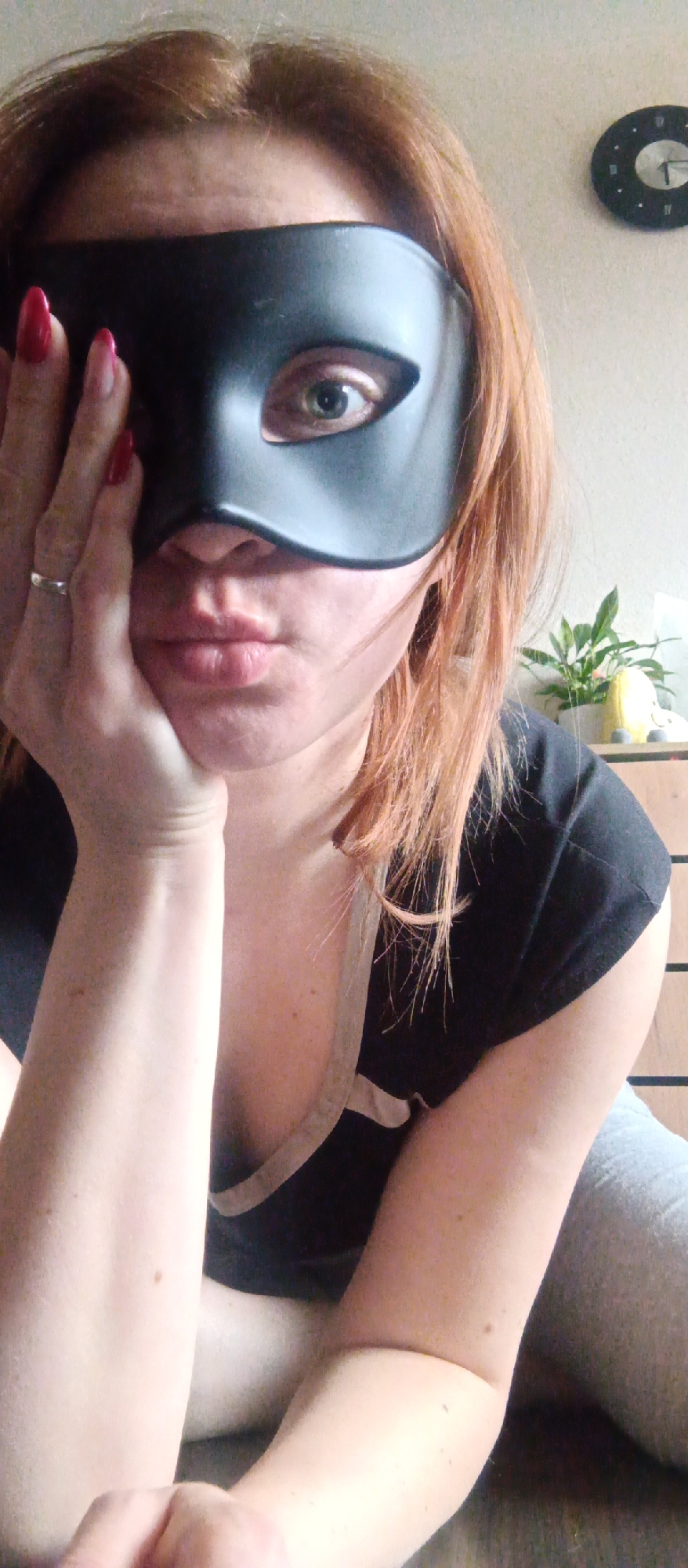 The image features an adult female in an indoor setting, posing with a black masquerade mask covering the upper half of her face. Her reddish-brown hair frames her face, and a striking green or hazel eye is visible through the mask. Her left hand, adorned with bright red nail polish and a silver-colored ring on her ring finger, rests against her cheek and chin, conveying a thoughtful or contemplative expression. She is wearing a black t-shirt with a light-colored trim and appears to be wearing grey casual pants or leggings.
The background shows a light-colored wall. A round, black wall clock with light-colored hands is visible in the upper right, indicating approximately 1:55. Below the clock, a small potted plant sits on a light brown wooden dresser or shelf. The lighting suggests it is daytime. There is no visible text that can be clearly read.