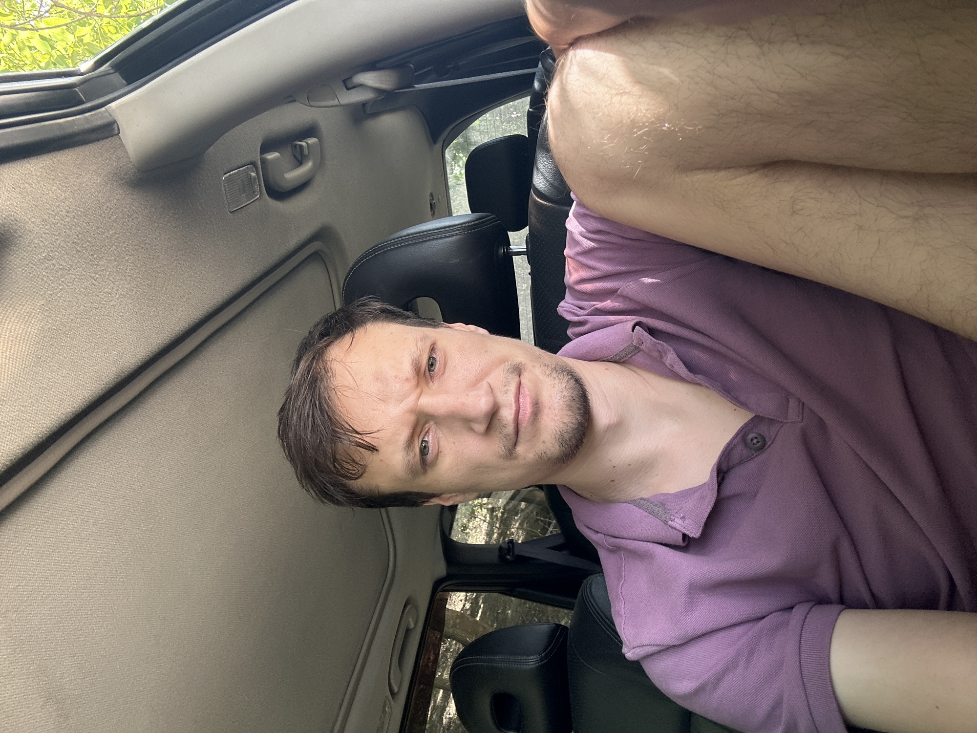 A man is sitting in the driver's seat of a car, which is parked outdoors in a wooded area near Makiv, Ukraine. The man is in his late 20s or early 30s, with light brown hair, a fair complexion, and stubble on his chin. He is wearing a purple polo shirt and has his right arm resting on his lap. The interior of the car, including the dashboard, ceiling, and seats, is visible. The sun is shining through the windows, creating shadows and highlights. Trees and foliage can be seen through the car windows, indicating an outdoor, natural setting.