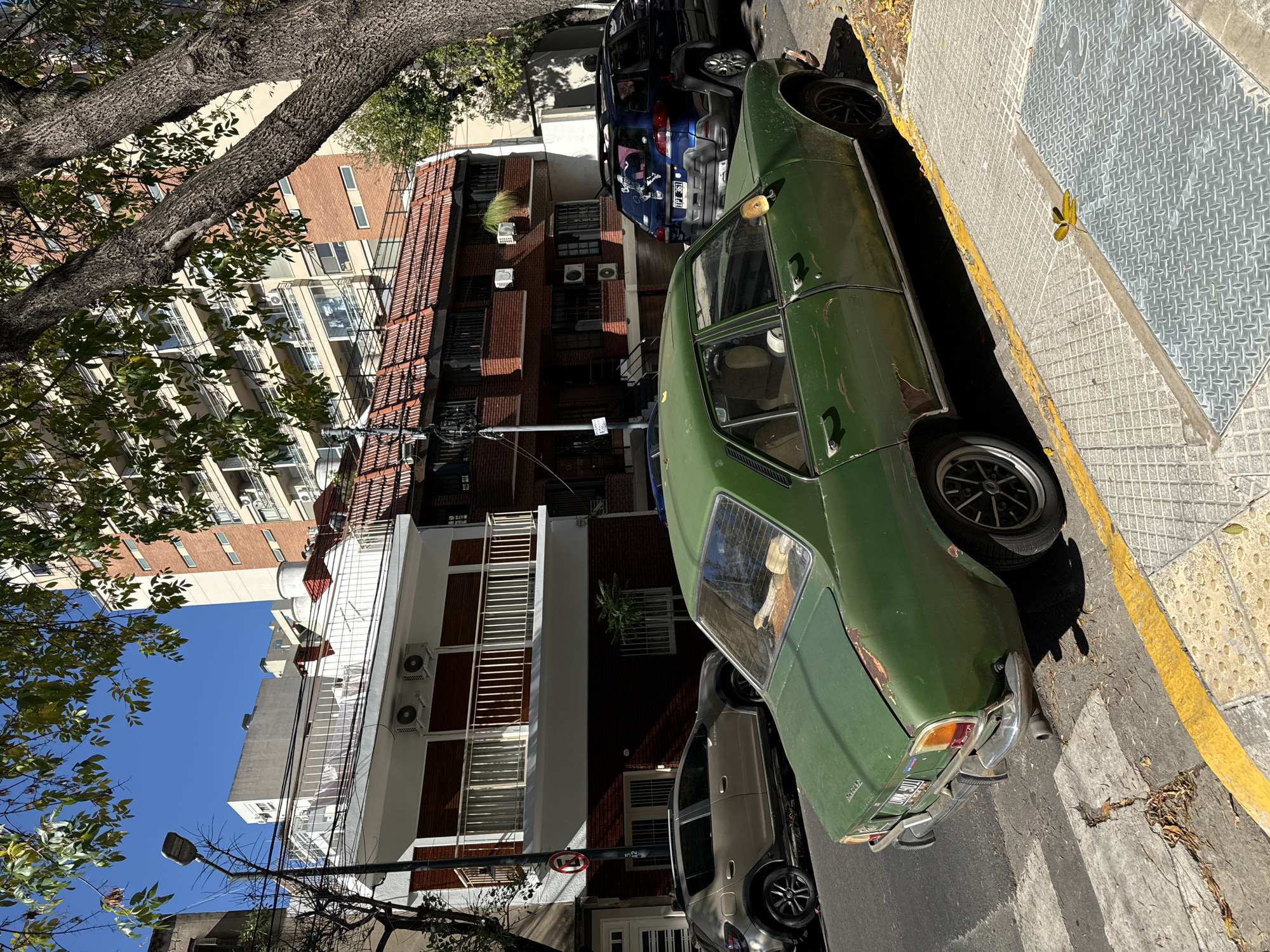 An eye-level shot of a street scene in Buenos Aires, Argentina, on a sunny day with a clear blue sky. An old, dark green car, parked facing left, dominates the foreground. Behind it, a compact silver car is parked. A blue SUV is visible further back, also parked. Cars are parked tightly along the curbside.
Behind the cars, buildings of varying heights and architectural styles are visible. Some are brick with dark red tones and balconies. Others are modern, with white facades and many windows and air conditioning units. Tall trees, with a thick trunk and leaves, partially obstruct the view of the building tops. The sidewalk is made of grey concrete tiles. The edge of the sidewalk is marked with a yellow line. A scattering of leaves are on the street and sidewalk. A lamppost is visible on the left side of the image. The license plate of the green car has some obscured text. The license plate of the blue SUV reads "IPA 261." A "No Stopping" sign is next to the compact car.
