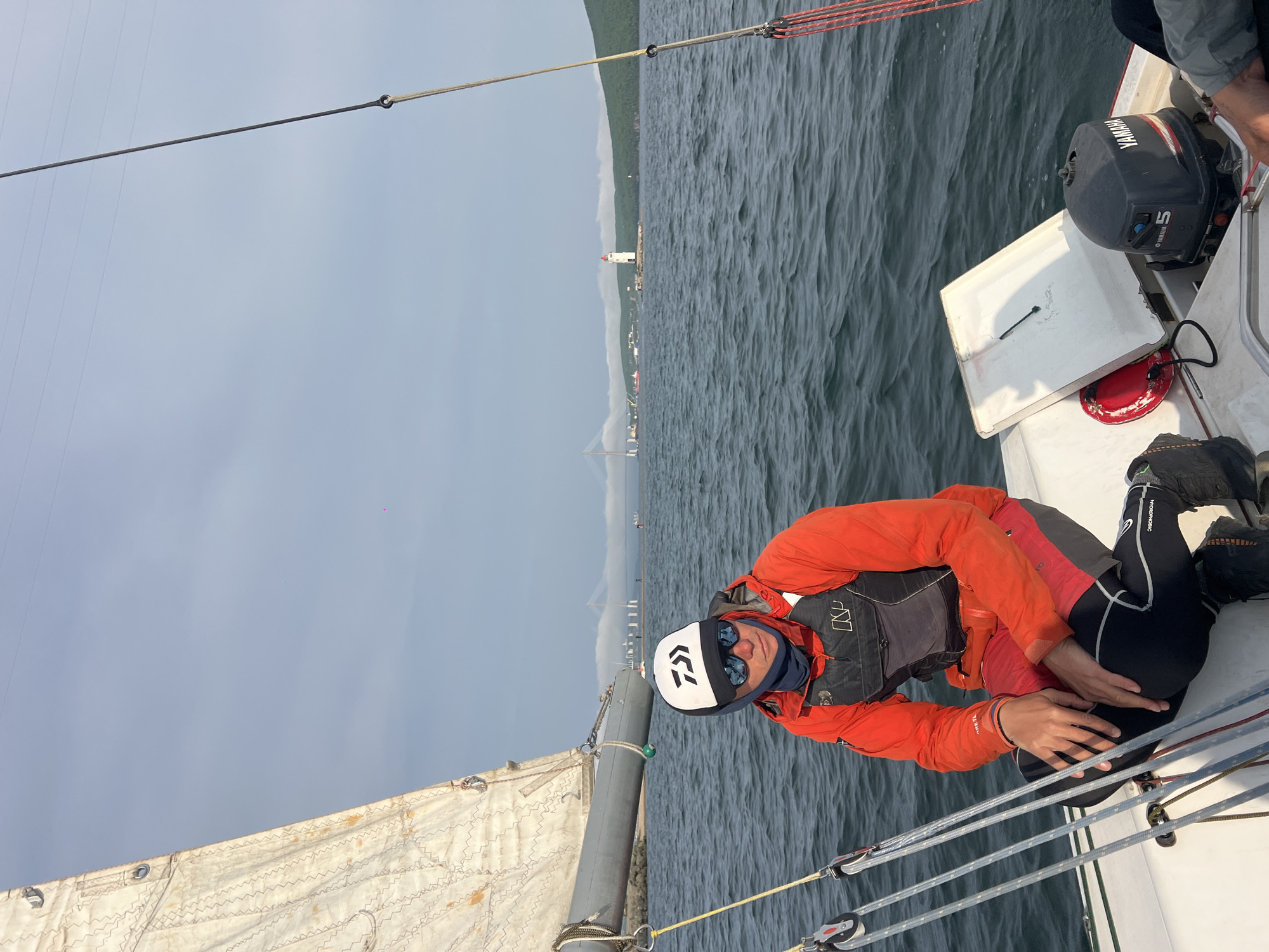 A man in an orange jacket, black and grey life vest, black leggings, and a white and black cap is seated on a sailboat. He is wearing sunglasses and has his hands positioned near his leg. The boat is on the water, and in the background is a view of Vladivostok, Russia, including a bridge and a lighthouse on the shore. An outboard motor labeled "YAMAHA 5" is attached to the boat behind the man. The sail is visible, along with ropes and rigging. The sky is overcast.