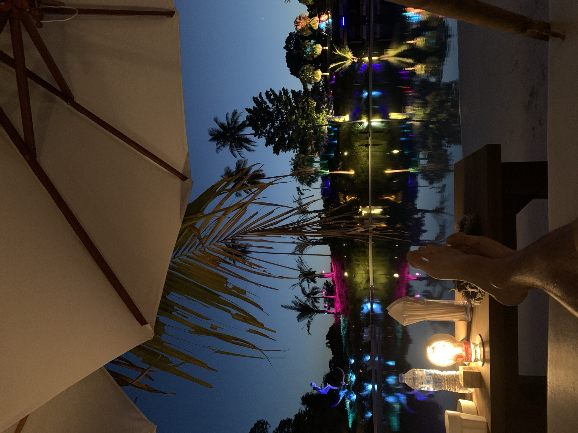 The image shows an evening view in Baan Lipa Noi, Thailand, taken from a relaxed, seated position. An open white patio umbrella is partially visible in the left foreground. Tropical palm fronds reach across the lower left portion of the frame toward the twilight sky. A body of water reflects colorful lights from illuminated trees and structures in the distance, suggesting a resort or landscaped area. Near the right edge of the frame are a person's feet resting on a surface, alongside a small lamp, a water bottle, and other items, indicating a comfortable setting.