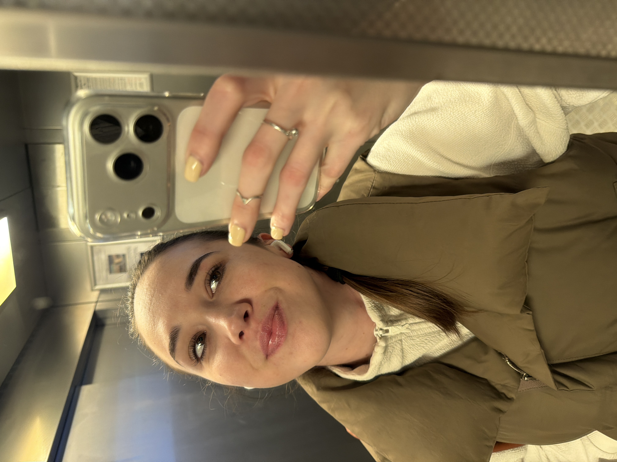 A woman takes a selfie in what appears to be an elevator. She is wearing a tan puffer vest over a white zip-up hoodie. She is holding a phone with three camera lenses in her right hand, her nails are painted a light yellow. The background shows the interior of an elevator with a grey metal finish. The image includes a reflection of the elevator interior. There is no definitive evidence to suggest this image was taken in Rostov-on-Don, Russia.