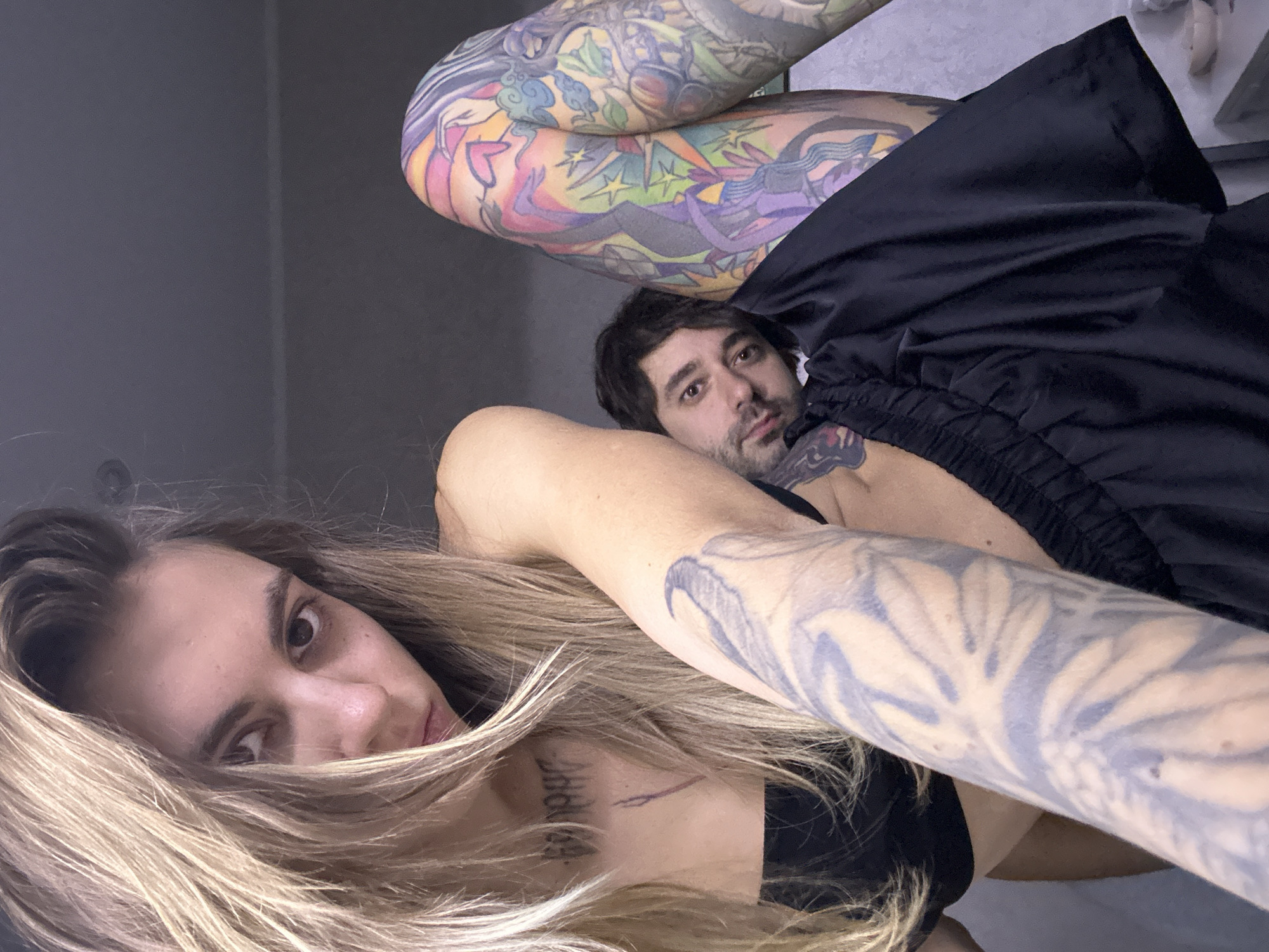 The image shows two individuals, a woman and a man, in close proximity. The woman has long blonde hair, and is wearing a black top. Her left arm is tattooed and extends across the frame. The man, with dark hair and a beard, is looking towards the camera from behind the woman's arm. He is partially covered by a black fabric, possibly clothing. The background is a plain, light-colored wall. The overall lighting is soft, suggesting an indoor setting. No specific location in Moscow, Russia, is discernible from the image.
