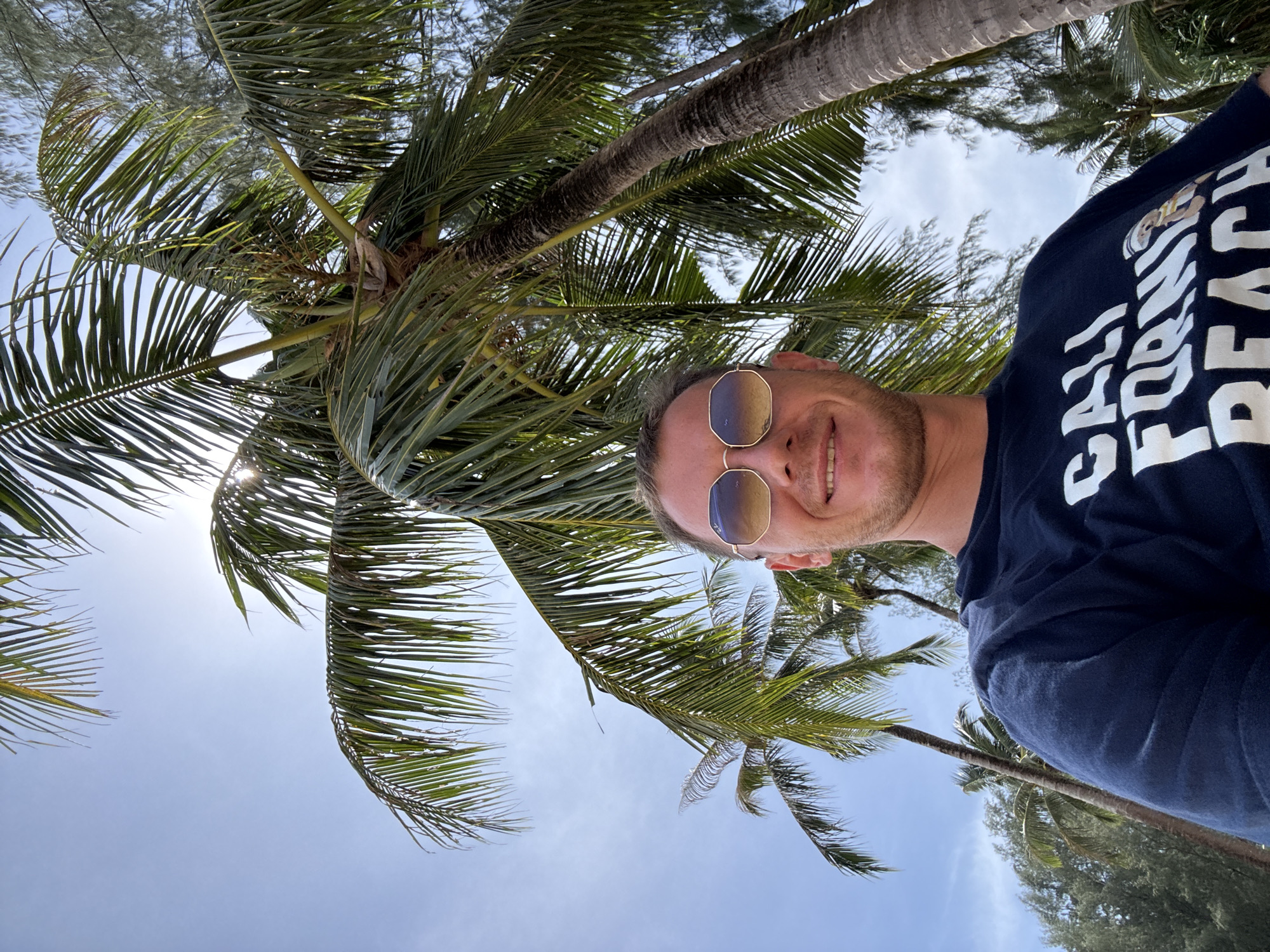 A man wearing sunglasses and a dark blue t-shirt with white lettering stands outdoors under palm trees. The man is smiling and looking at the camera. The t-shirt has the words "CALI FORNI BEACH" printed on it, along with an image of a bear wearing a sailor's hat. The background consists of palm trees and a bright blue sky. The location is Ban Khuk Khak, Thailand.