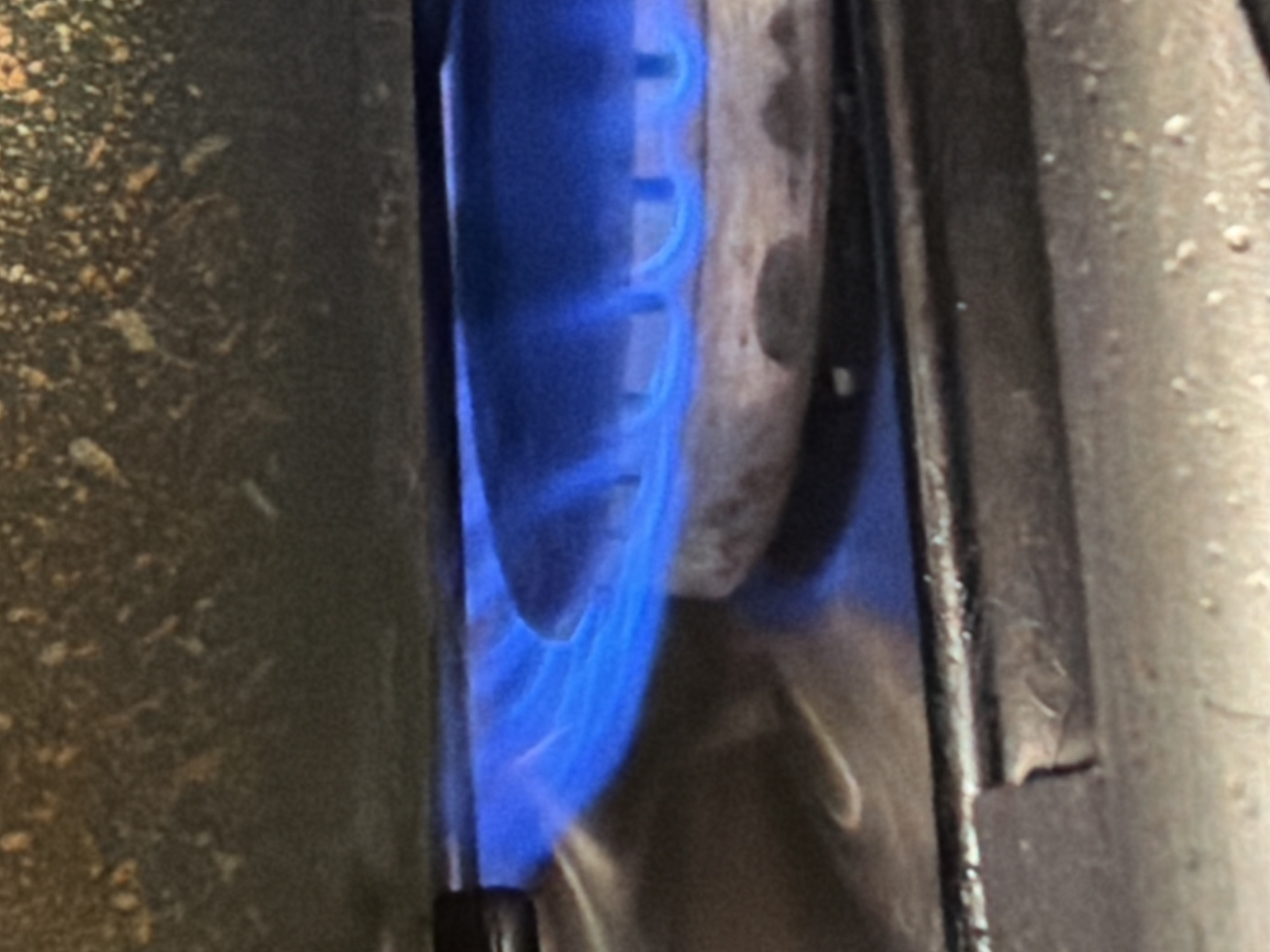 A close-up, medium shot captures a gas burner on a stove with a bright blue flame. The flame is emanating from a circular burner with several small holes from which the gas is escaping. The burner is positioned on a metallic surface, and above it, a dark, grimy grate or a part of the stove's interior is visible. The image is well-lit, focusing on the intense heat and light of the gas flame. The scene appears to be indoors, likely a kitchen, and there are no people or other discernible objects in the frame, emphasizing the visual of the burning gas. The overall impression is one of controlled combustion and the operation of a common kitchen appliance. It is not possible to determine the specific location within the city of Khimki, Russia, based on this image alone, as it lacks any identifying landmarks or context.
