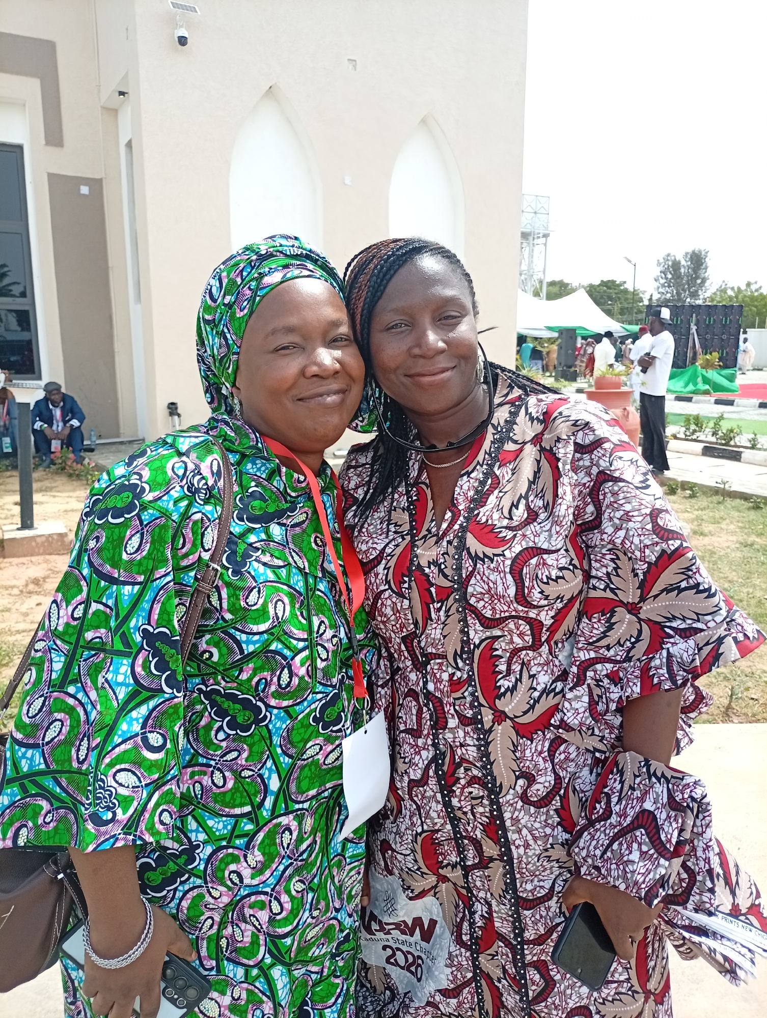 Two women, dressed in vibrant traditional African print attire, pose together and smile at the camera outdoors. The woman on the left wears a green, blue, and pink patterned headscarf and a matching dress. She has an ID lanyard with a white card around her neck, a brown shoulder bag, and is holding a black smartphone with a silver camera module. The woman on the right wears a red, brown, and white patterned dress, with her hair in braids and sunglasses perched on her head. She also has an ID lanyard, part of which is visible.
They appear to be at an outdoor event during bright daylight, suggested by the clear skies and shadows. In the background, there is a light-colored building with arched windows or doorways, possibly a mosque or public building, equipped with a security camera and a small solar panel. Several other people are visible in the distance, some seated and some standing near white and green tents, indicating a gathering or event. Sound equipment, possibly speakers or screens, can also be seen. The setting includes manicured green spaces and paved areas, suggesting an organized venue.
Visible text on the woman on the left's lanyard or a document held in her hand includes "KADUNA," "2026," and "PRINTS NEW CHICA." On the woman on the right's lanyard, "KADUNA STATE CHAPTER," and "2026" are partially legible, along with an acronym that could be "RW" or "RNW." Both women exhibit warm smiles, conveying a friendly and joyful mood. The attire and general setting are consistent with the provided location context of Unguwan Sanusi, Nigeria.