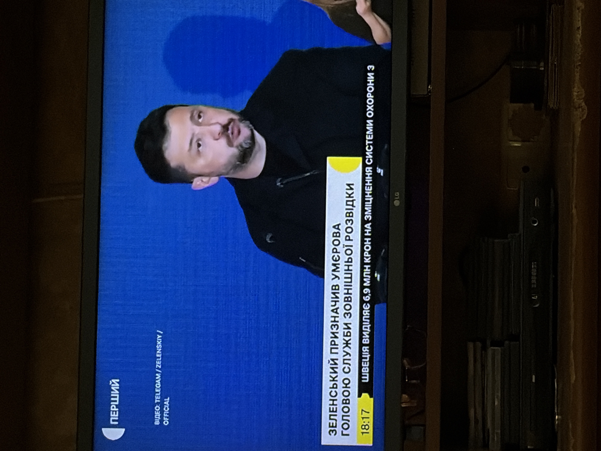 The image shows a television screen displaying a news broadcast. In the foreground, a man, presumably a news anchor or interviewee, is visible from the chest up. He is wearing a dark jacket and has short, dark hair and a beard. The background is a solid blue screen with some subtle shadowing. A news ticker runs along the bottom of the screen, displaying text in Ukrainian. The text at the top of the screen indicates "ПЕРШИЙ," which is likely the name of the television channel. Below that, it reads "ВІДЕО: TELEGAM / ZELENSKIY / OFFICIAL." The ticker displays "18:17" followed by Ukrainian text. The television itself is mounted on a dark wooden surface, and below it, stacks of CDs or DVDs and a black electronic device are visible. The overall lighting suggests an indoor setting, likely a living room or entertainment area.