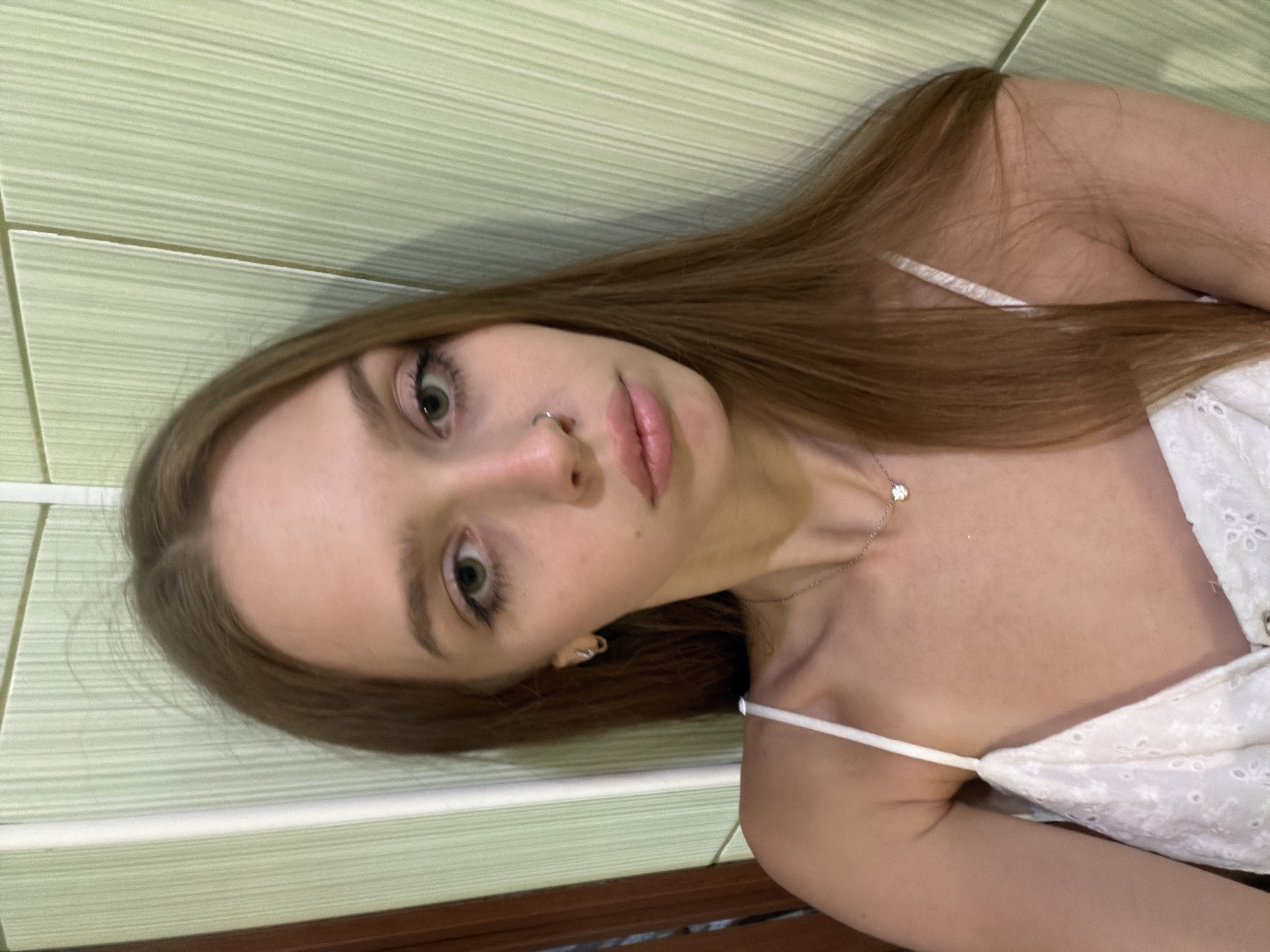 This is a close-up, vertically oriented selfie of a young woman with long, light brown hair. She is wearing a white top with thin straps and what appears to be delicate embroidery. A thin silver necklace with a small, sparkling pendant rests on her collarbone. Her left eye is visible, open and looking towards the camera, with long eyelashes. She has a subtle nose ring. Her lips are slightly parted, and her skin appears fair with a hint of blush. The background consists of light green, horizontally striped wall tiles and a dark brown surface at the bottom, possibly a piece of furniture. The lighting suggests an indoor setting, possibly during the day, as there are no strong shadows. The overall mood of the image is casual and relaxed.