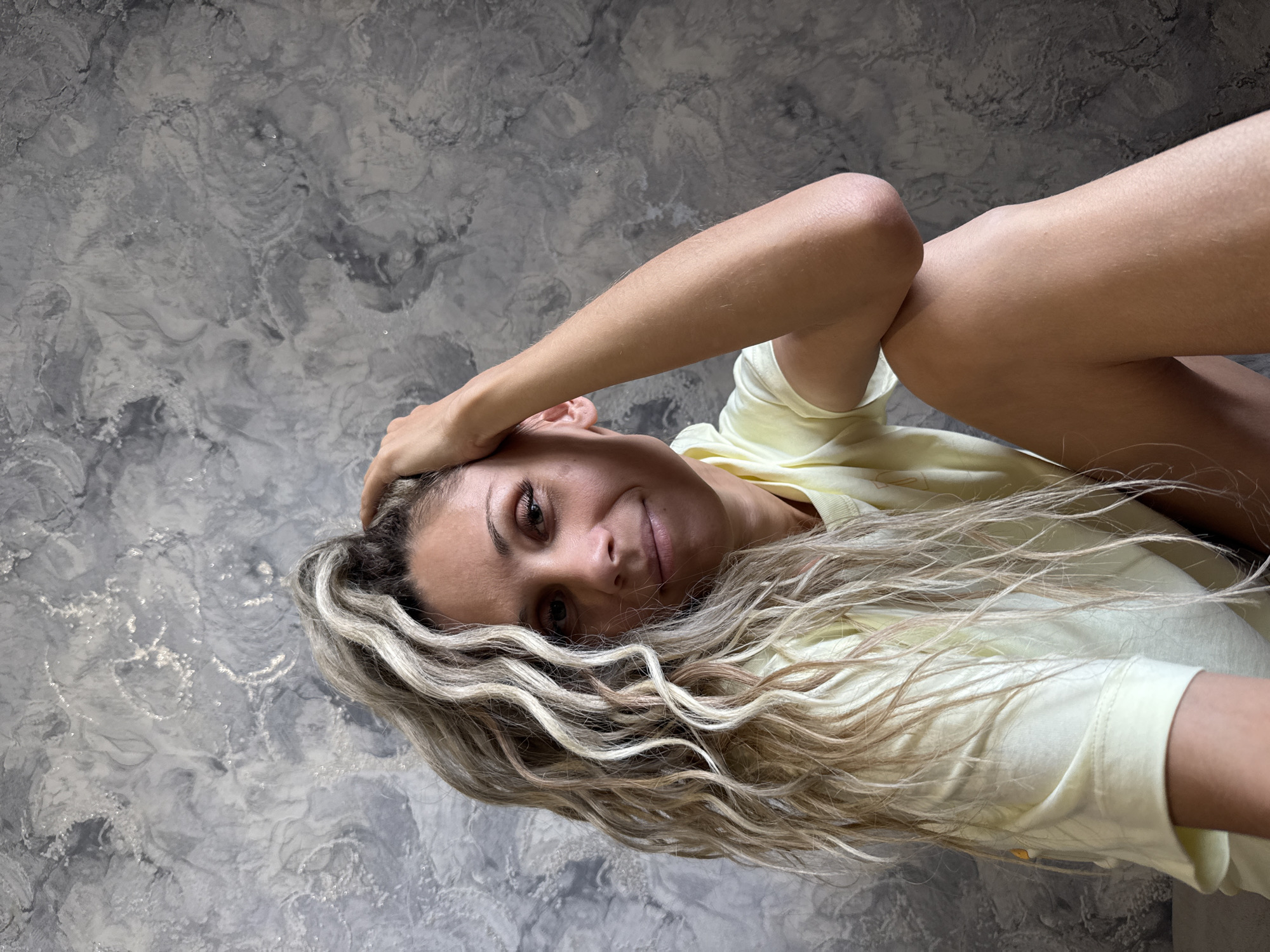 In Rusanovo, Russia, a woman with long, wavy blonde and light brown hair is seated in front of a textured grey wall. She is wearing a pale yellow t-shirt and has her right arm bent, with her hand resting on her head. Her left leg is bent at the knee and crossed over her right leg, with her right arm resting on her left knee. The woman is looking towards the camera with a slight smile. The background is a grey, marbled textured wall.