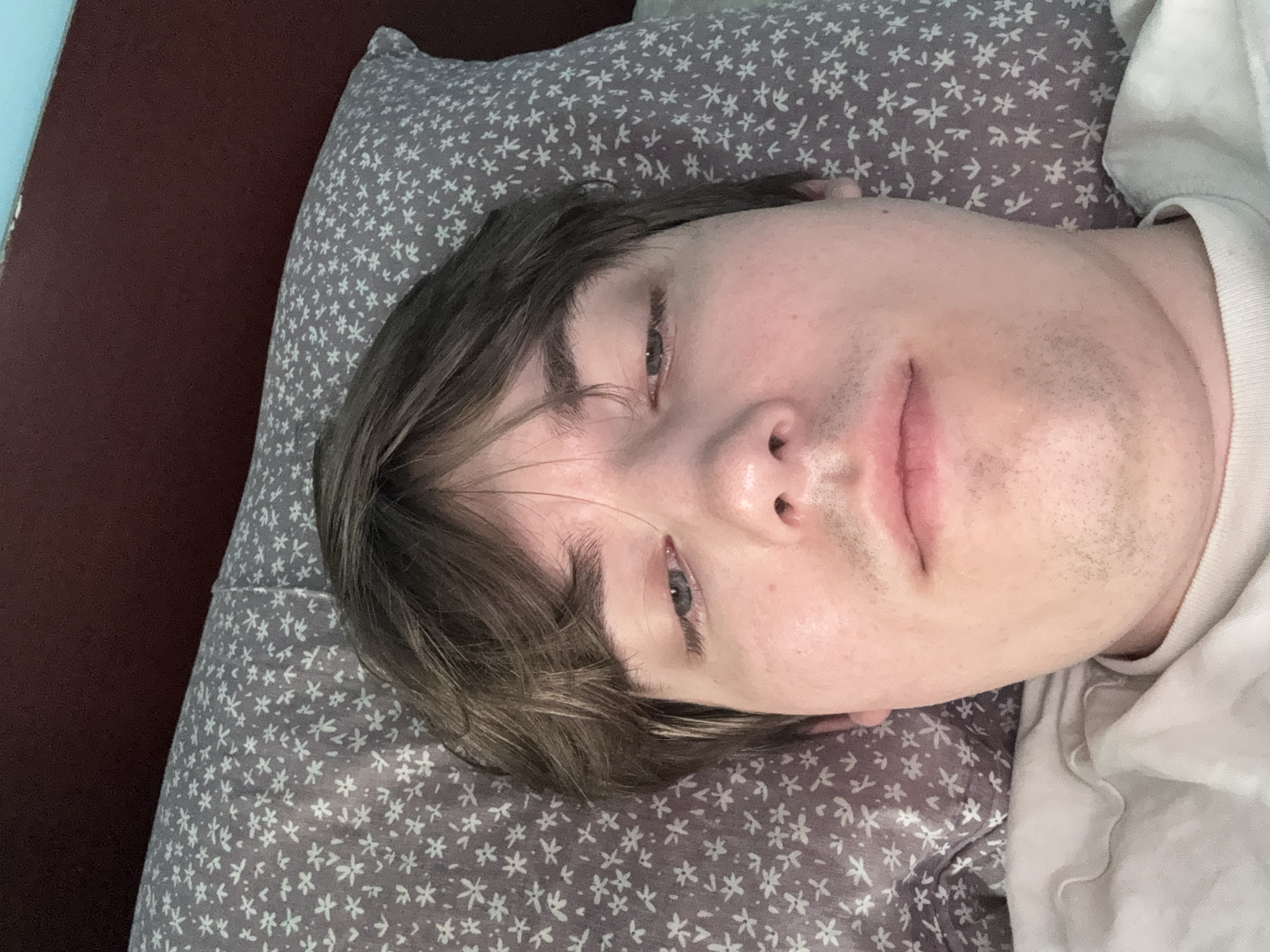 A close-up, overhead shot shows a young man lying on a floral patterned pillow in a bed. The man's face fills the majority of the frame, with his eyes partially closed and his gaze directed downward. He has brown hair, light skin, and stubble on his chin and upper lip. He is wearing a white t-shirt. The background consists of a dark headboard and a sliver of a light blue wall. The scene appears to be indoors, in a bedroom setting, but the specific location in Dnipro, Ukraine, cannot be determined from the provided image.