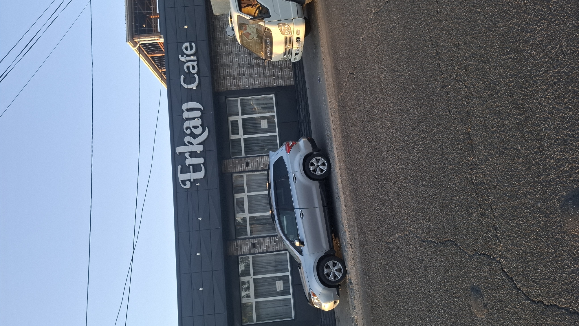 The image shows the facade of a building with the sign "Erkan Cafe" in white lettering. The building has a dark grey exterior and large windows with white curtains. In front of the building, there is a silver SUV parked on the sidewalk and a white truck parked on the street. The street in the foreground is asphalt with visible cracks. The sky is clear and blue. The building is located on a street in Shymkent, Kazakhstan.