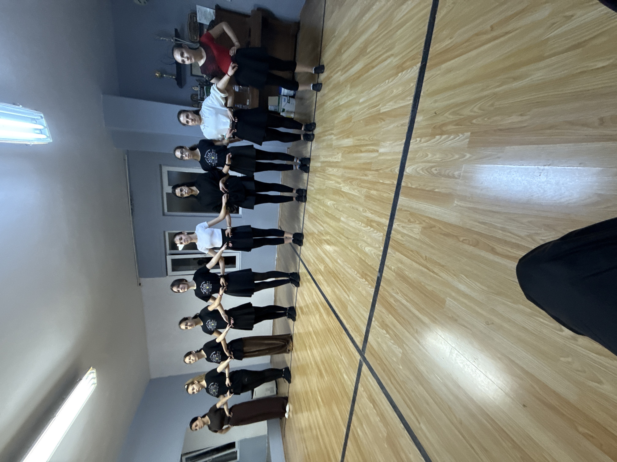 Eleven young women are captured in an indoor setting, likely a dance studio or practice room, in Buhovo, Bulgaria. The image, rotated counter-clockwise, shows them standing in a linear formation, facing the viewer. They are arranged shoulder-to-shoulder, seemingly connected by holding hands or resting hands on each other's arms, forming a unified group.
The majority of the women are dressed in black tops and black skirts or pants, with several wearing black shoes, suggesting a uniform or practice attire. Variations in clothing include one woman at the front wearing a red short-sleeved top with black bottoms, another in a white short-sleeved top and black bottoms, and two women towards the back in brown skirts or pants paired with black tops. Their expressions are generally neutral or convey a slight smile, consistent with posing for a group photograph.
The room features a polished light wooden floor with a prominent black dividing line extending across it. The walls are a light grey color, and the ceiling is equipped with several long, rectangular fluorescent light fixtures that illuminate the space. In the background, behind the women on the right side, a dark wooden cabinet or shelf holds a few indistinct objects, possibly trophies or decorative items. The artificial lighting indicates that the photo was taken indoors, possibly during the evening or night. No legible text is visible on any clothing or in the surroundings.