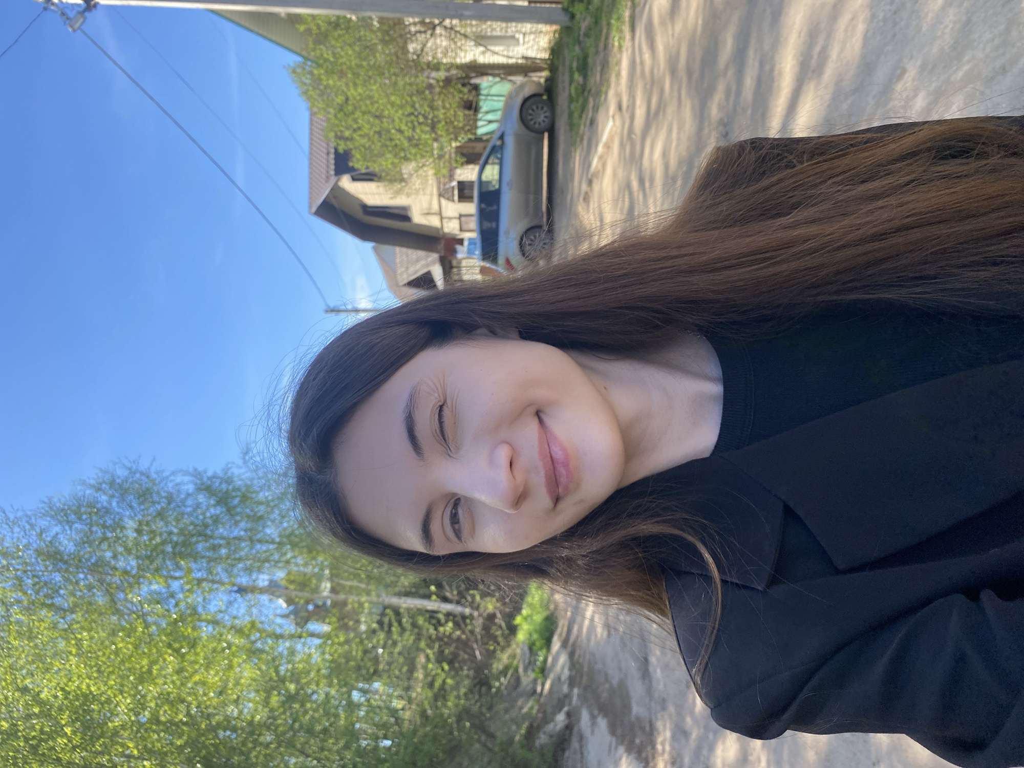 A woman is centered in the foreground, winking at the camera. She has long brown hair and is wearing a black top and jacket. The background shows a residential area on a sunny day. There is a house with a brown roof and a light gray car parked in front. Trees with green leaves line the street. The location is likely in Votkinsk, Russia, based on the question's premise.