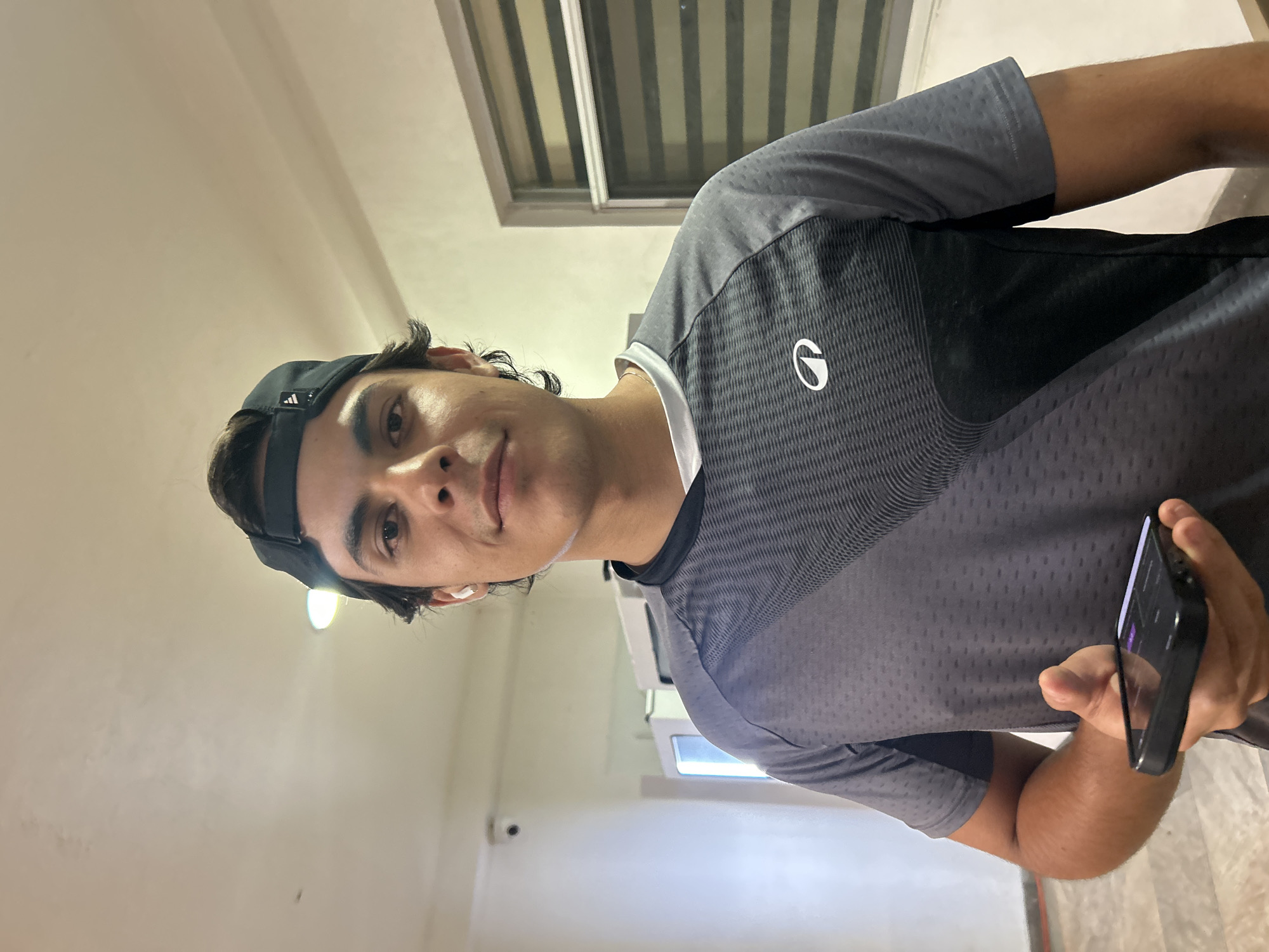 A person wearing a black baseball cap and a gray and black shirt is holding a black smartphone in their right hand. The person is indoors, possibly in a hallway or office space. A window with blinds is visible in the background. The location appears to be Unknown, Unknown.