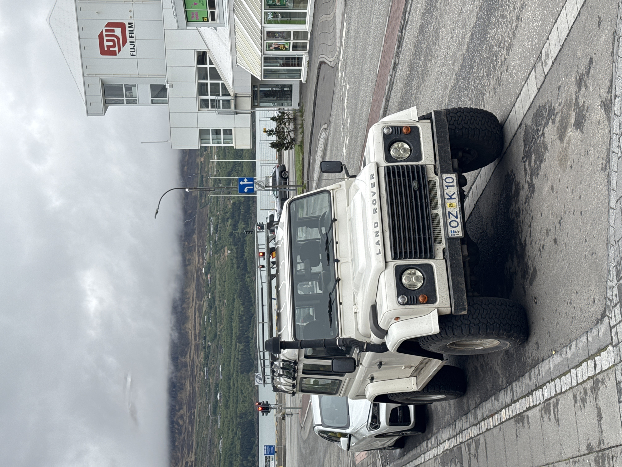 The image, oriented on its side, captures a street scene in Akureyri, Iceland, centered on a white Land Rover Defender 110. The rugged off-road vehicle features prominent modifications including large knobby tires, a snorkel extending up the passenger side, and a robust roof rack with multiple auxiliary lights. The words "LAND ROVER" are clearly visible on the vehicle's front grille. Its Icelandic license plate, "OZ K 10", bears the country code "IS". A portion of a newer white SUV is visible parked behind the Defender.
In the mid-ground, a modern white building stands tall, displaying a large red "FUJIFILM" logo and text. Adjacent to it, another building features a green sign with the word "Optic". The street is a mix of asphalt and cobblestones, appearing damp, indicative of overcast or recent rainy weather. Traffic lights, a street lamp, and a blue road sign indicating options to "turn left or go straight" are also present. A small, partially visible "LEXUS" sign is affixed to a pole near the traffic lights.
The background reveals a dramatic landscape of green, forested areas and mountains partially enveloped in dense, low-lying clouds or fog, creating a moody, overcast atmosphere. The sky is uniformly gray. The scene conveys a blend of urban infrastructure and the wild, natural environment characteristic of Iceland, with the durable Land Rover fitting well into this context.