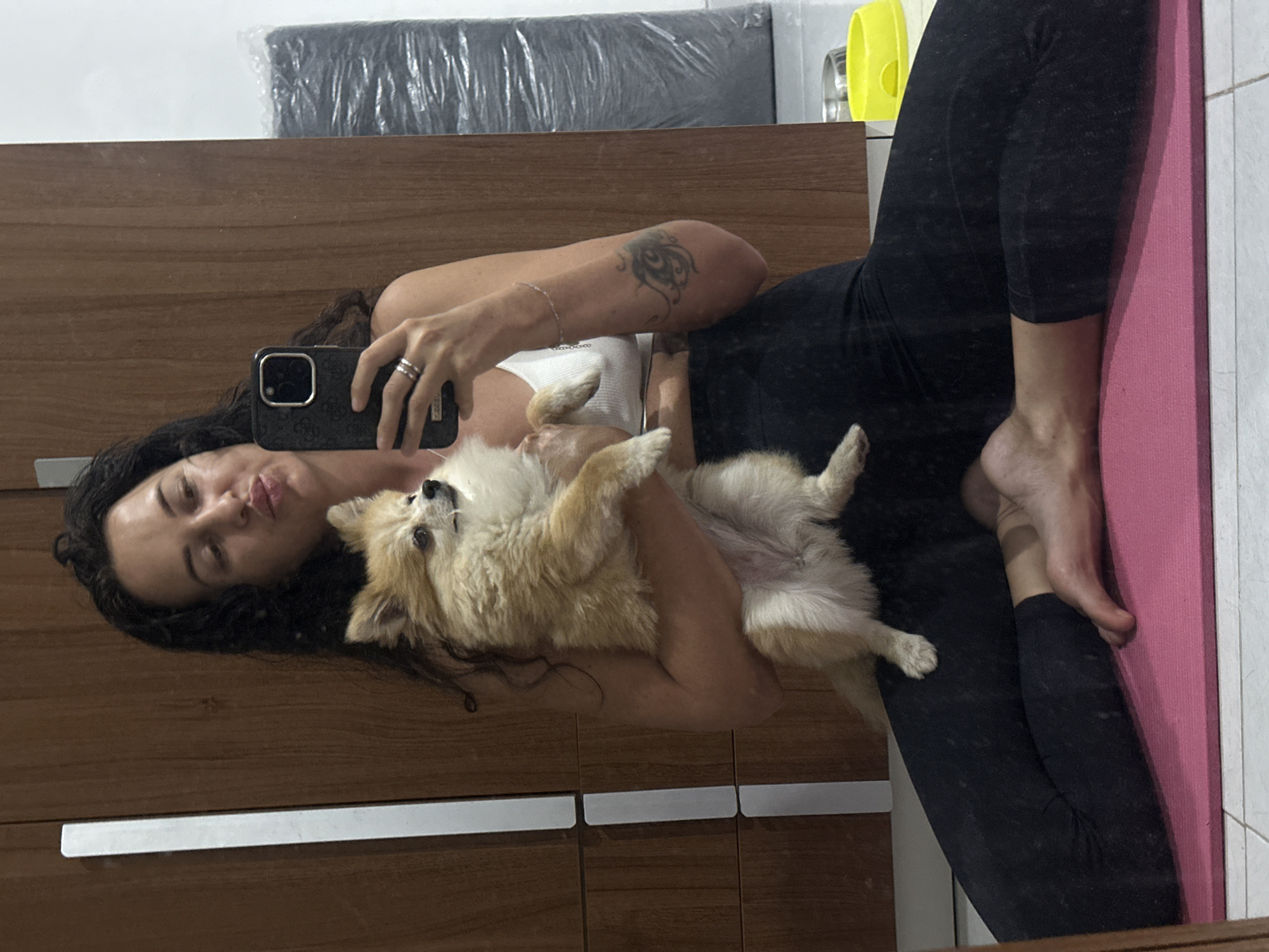 A woman is taking a mirror selfie with a small tan and white dog in her arms. She is wearing a white tank top and black leggings, and is sitting on a pink yoga mat. The dog is looking to the side, while the woman is puckering her lips. In the background, there is a wooden closet and a rolled-up black object. There is also a yellow dog bowl and a metal dog bowl visible. The scene appears to be indoors, possibly in a bedroom or living room, in Kuta Selatan, Indonesia.