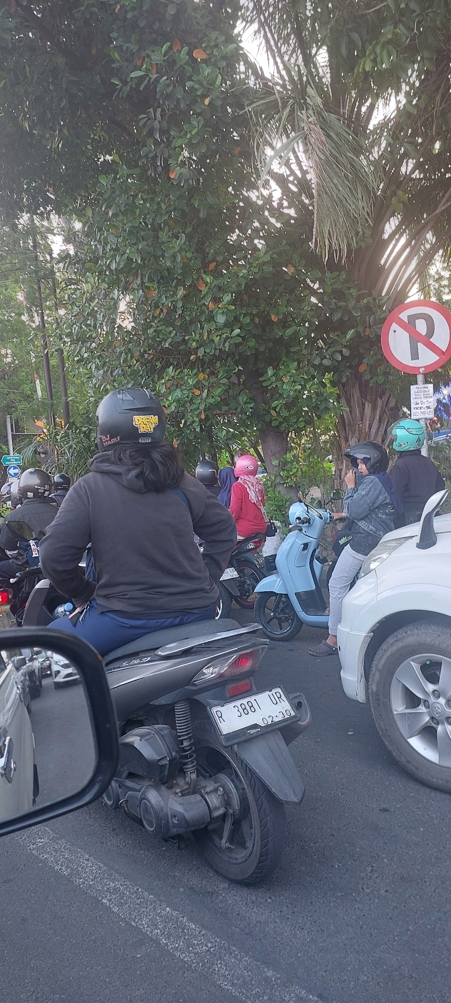 The image shows a street scene in Surabaya, Indonesia, with several motorbikes and a car. The focus is on a person riding a grey scooter in the foreground, wearing a black hoodie and a black helmet with yellow decals. The scooter has a license plate reading "R 3881 UR". In the background, there are other motorbikes with riders wearing helmets of various colors, including pink and green. A white car is parked to the right of the scooter. Trees and foliage are visible, along with a "No Parking" sign. The scene appears to be during the day, with overcast skies.