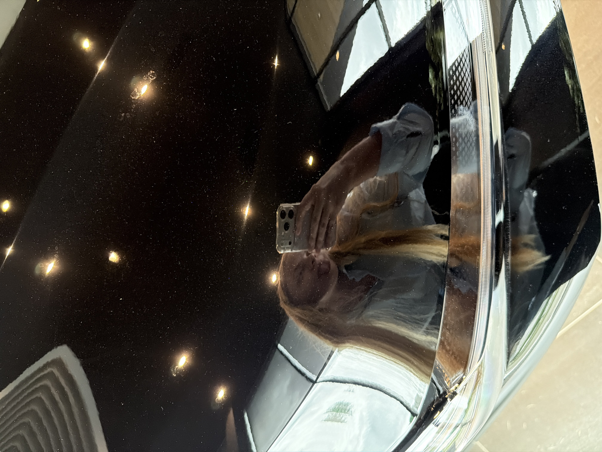 The image shows a reflection in the shiny black surface of a car, presumably in Izhevsk, Russia. The reflection includes a person taking a picture with a smartphone. The reflection also shows windows and a building facade. The right side of the frame contains the edge of a car headlight. The black surface has numerous small, bright spots, likely light reflections or imperfections.