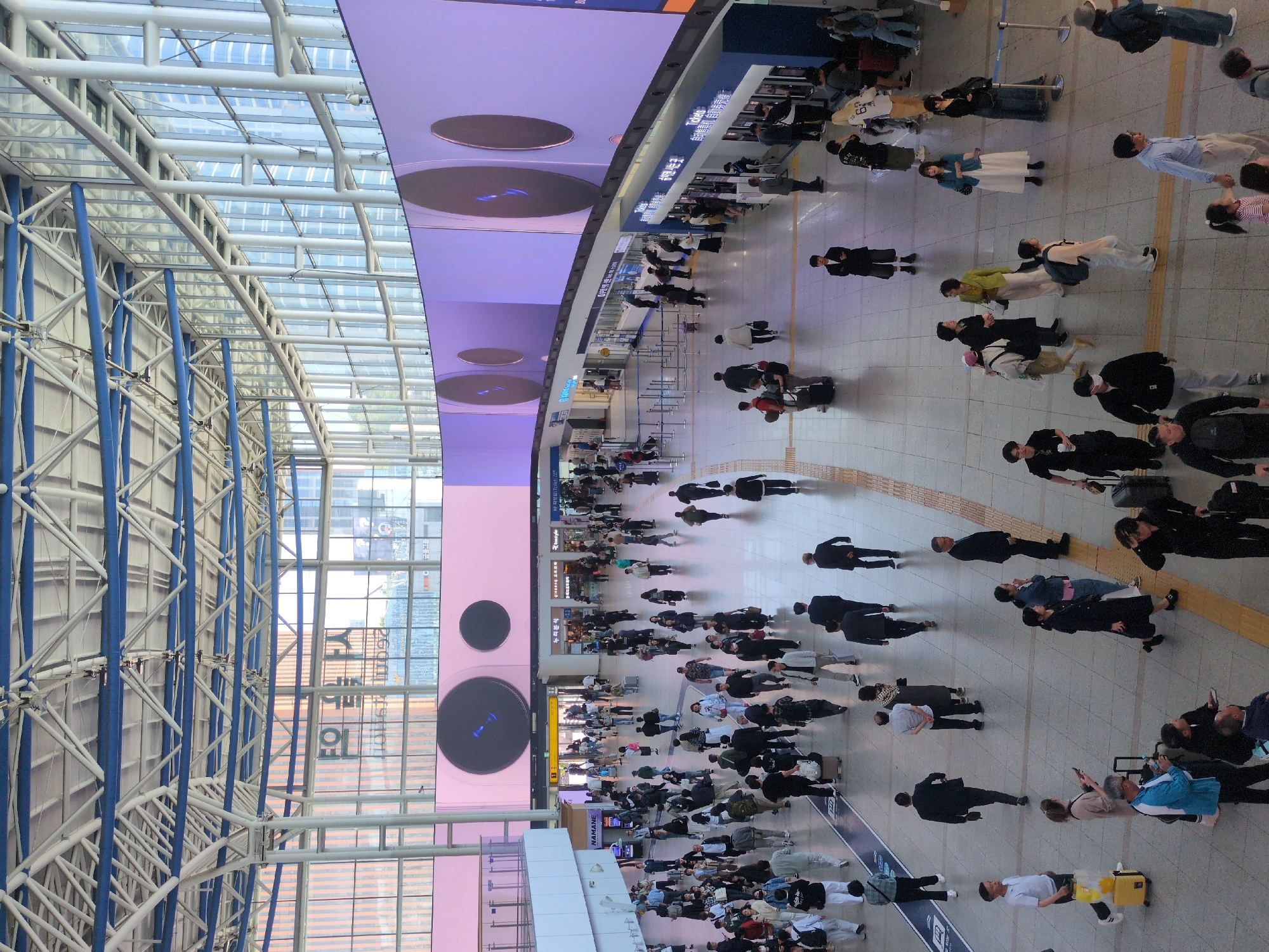 This is an aerial view of a bustling indoor space, likely a train station or transportation hub, in Seoul, South Korea.
The building has a high, arched roof supported by white steel beams, some of which are reinforced with blue. Natural light streams in through a glass section of the roof.
A large, rectangular digital display hangs above the main area, featuring a pink background with multiple circular screens.
Numerous people are walking around in various directions, predominantly dressed in dark clothing. Some are pulling luggage or using smartphones. The floor is tiled in a neutral color, with yellow tactile paving.
There are ticket counters in one area, with people queueing. The overall atmosphere is one of movement and activity.