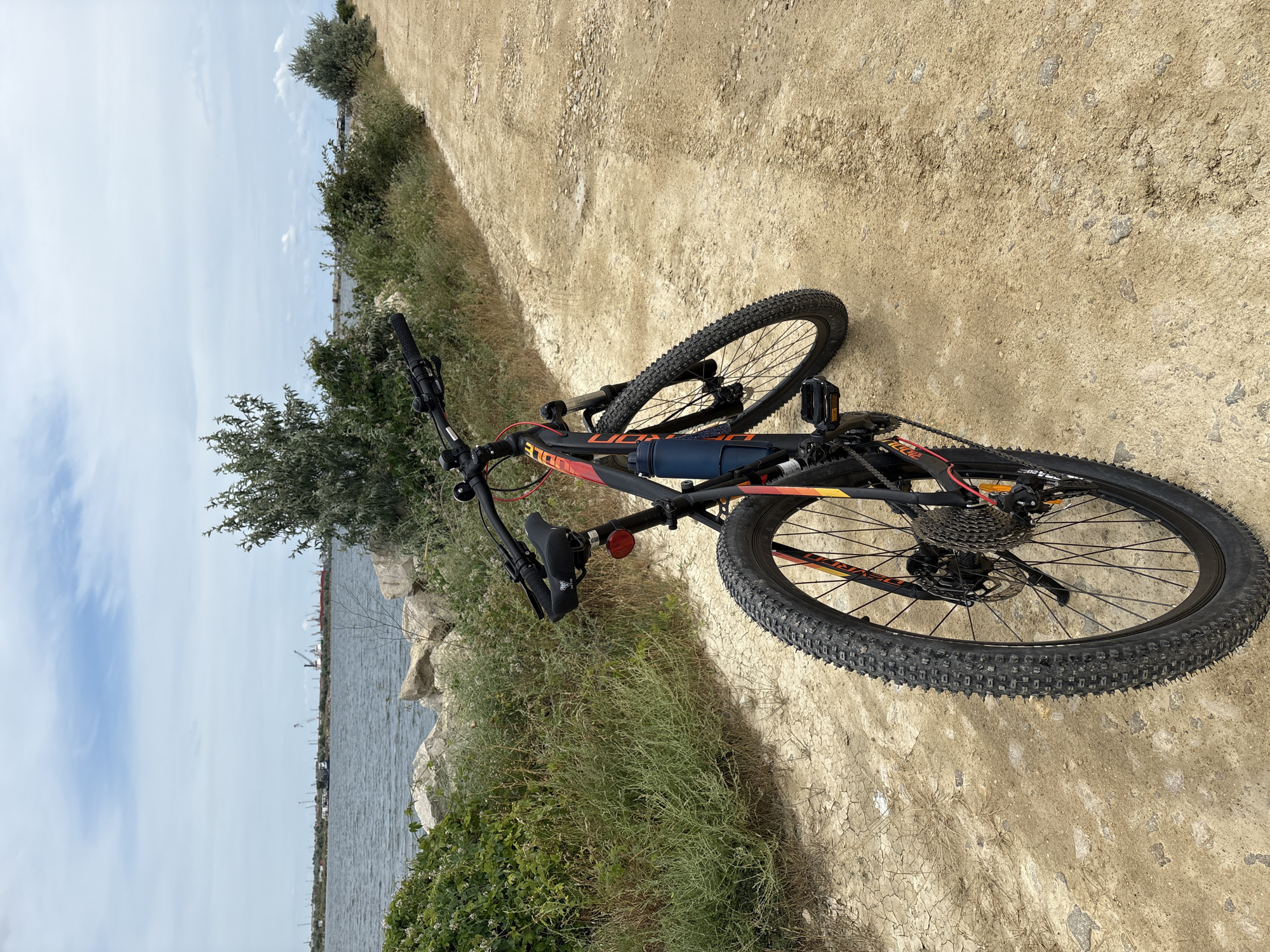 The image depicts a mountain bike parked on a dirt path next to a body of water. The bike is black with red and yellow accents and has a blue water bottle attached to its frame. The path is dry and dusty, with some loose rocks. In the background, the water stretches out to the horizon, with a cityscape featuring cranes and industrial structures visible in the distance. The sky is partly cloudy. The location is identified as the Năvodari City, Romania.