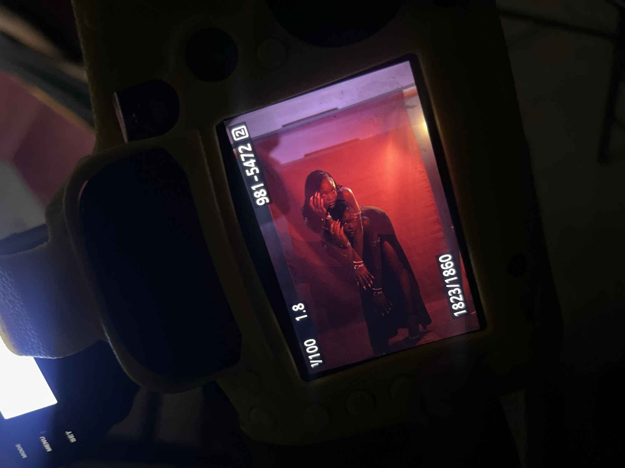 The image shows a close-up of a digital camera's LCD screen displaying a photograph. The camera is partially covered by a yellow silicone case. The photograph on the screen features a person with dark hair, wearing jewelry, against a red background. The camera's display shows technical information including "1/100 1.8", "981-5472 2", and "1823/1860". The buttons "MODE", "MENU", and "SET" are visible on the camera body. The scene is dark, with the camera's screen being the primary light source. The location in the city of Gura Top, Nigeria, cannot be determined from the provided image.
