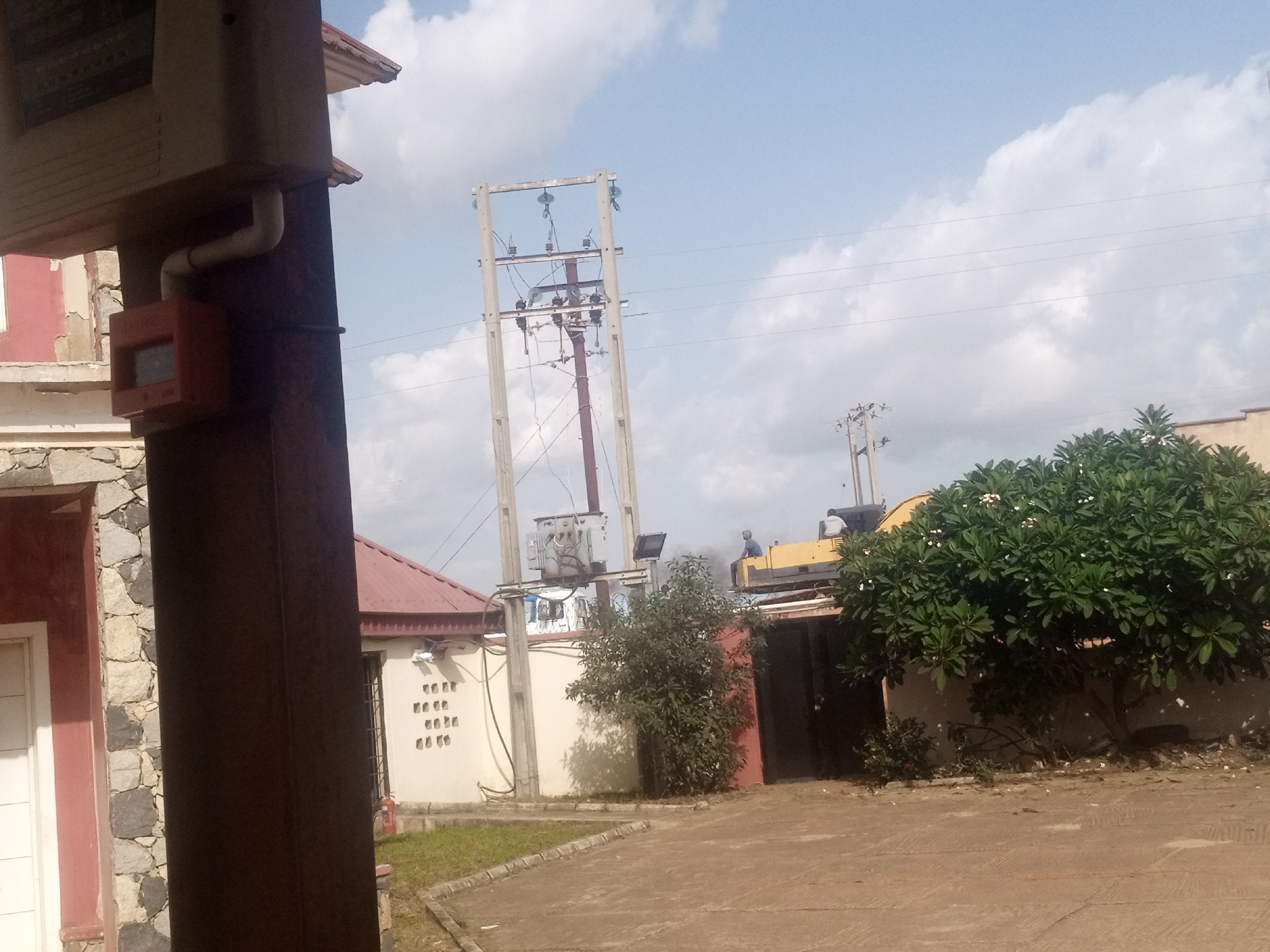 The image captures an outdoor scene with a focus on utility infrastructure and some surrounding buildings and vegetation. In the foreground and to the left, a large, dark brown vertical pillar is prominent, with a red electrical box attached to it. Behind this pillar, a light-colored building with stone accents and a red roof is partially visible.
The middle ground of the image features a prominent electricity pole with multiple crossbars and insulators, suggesting it's part of a power distribution network. A large electrical transformer is mounted on this pole. To the right of the electricity pole, a bright yellow piece of heavy machinery, possibly a loader or forklift, is visible. A person is seated on top of this machinery. Next to the machinery, a lush green tree with white flowers is in full bloom.
In the background, a bright blue sky with scattered white clouds indicates fair weather. The scene appears to be set in a developed area, possibly a commercial or industrial zone, given the presence of the heavy machinery and utility infrastructure. The overall impression is a bright, daytime scene, likely mid-morning or afternoon due to the quality of light and shadows. No visible text is clearly legible in the image.