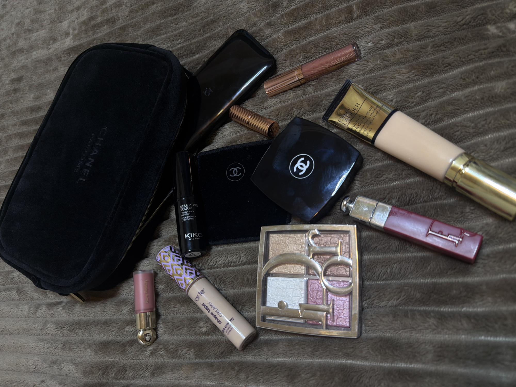 The image shows a collection of makeup products spread on a brown, textured surface. A black velvet Chanel makeup bag is positioned on the left. Various other products include lipsticks, lip glosses, foundation, concealer, a contour stick (Kiko Milano), an eyeshadow palette (Dior), a foundation tube (Estee Lauder), and a Tarte Shape Tape concealer. There is no information in the image about the city of Yerevan, Armenia.