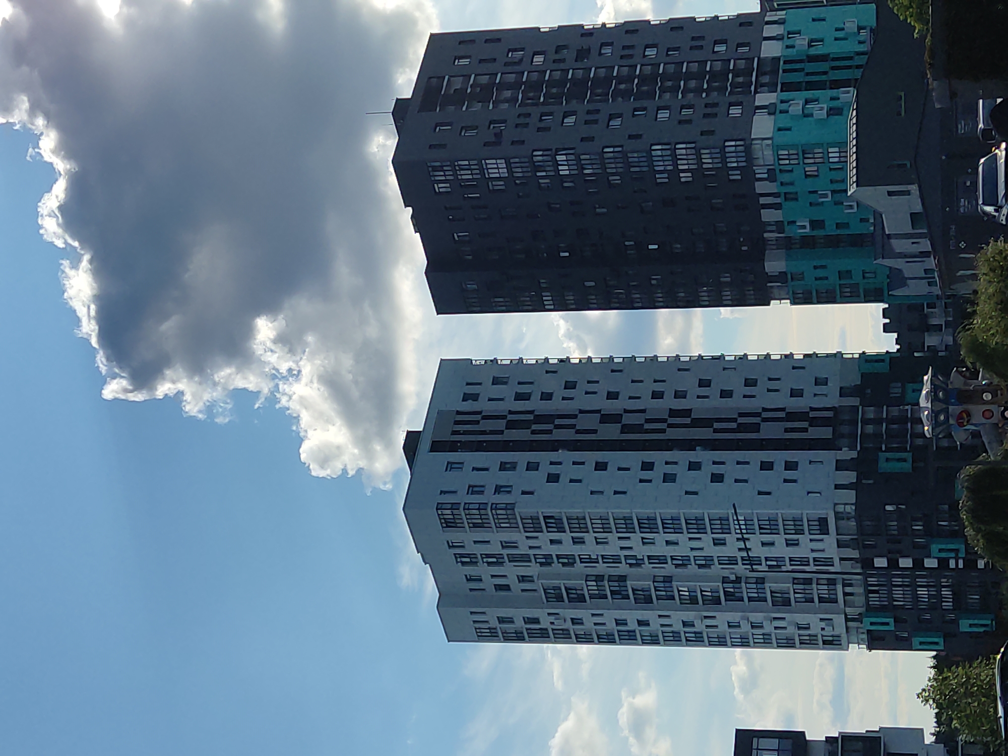 The image depicts a view of modern high-rise apartment buildings in the village council of Baravlyanski, Belarus. Two prominent buildings dominate the frame: one is a light-colored, multi-story structure with a grid-like pattern of windows and balconies, and the other is a dark-colored building with a similar window arrangement. The dark building features a section of bright turquoise colored panels towards its base, adding a vibrant contrast. The sky above is a mix of blue with scattered clouds, some of which are dense and appear to be illuminated by sunlight. In the foreground, the tops of trees and other smaller structures are visible, suggesting a developed urban or suburban landscape. The overall impression is one of contemporary architecture set against a dynamic sky.