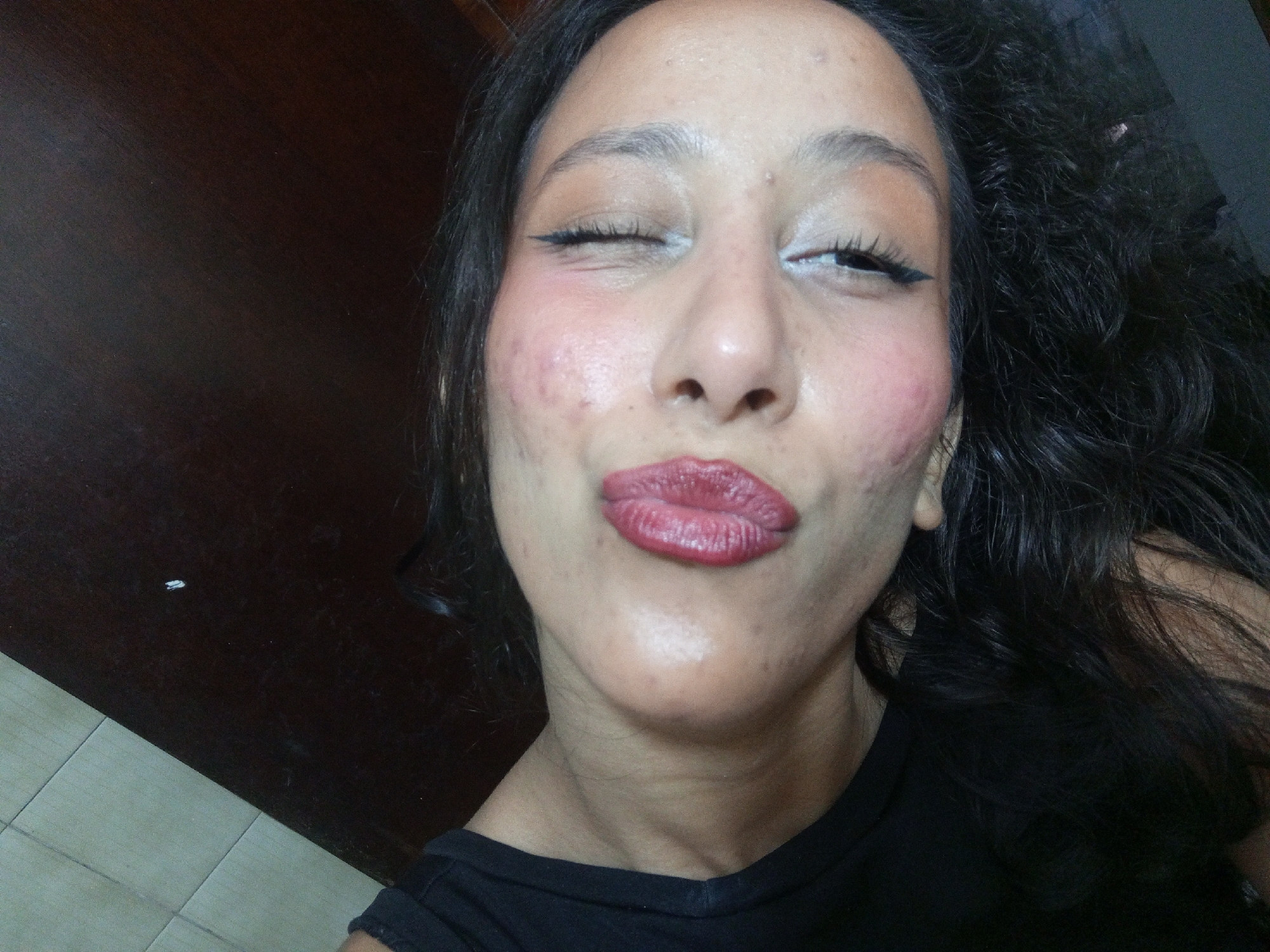 A person with dark hair is close to the camera, making a kissing face. The person is wearing a black top and makeup including eyeliner, eyeshadow, and lipstick. The background consists of a dark wooden surface and tiled floor. There is no indication of the location being Porlamar, Venezuela.