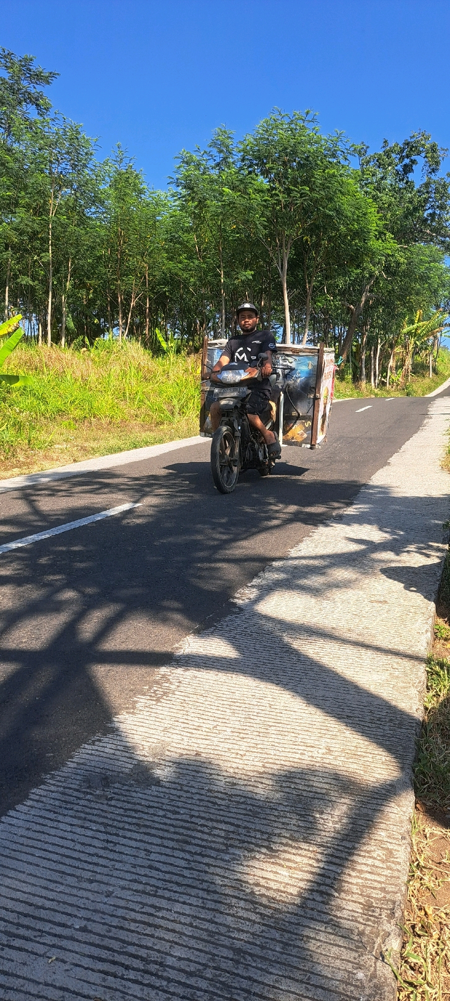 A man wearing a black t-shirt and shorts rides a motorcycle with a sidecar along a paved road in Sekarjoho, Indonesia. The motorcycle is black and has a license plate with the characters "B 2069 YDS". The sidecar is large and appears to be made of metal, with images of food items, possibly for a food business. The road is asphalt and has a white line marking the lane. There is a concrete shoulder on the right side of the road with grass growing along its edge. The background consists of lush green trees and vegetation, with a clear blue sky overhead. The scene is illuminated by bright sunlight, casting shadows on the road.