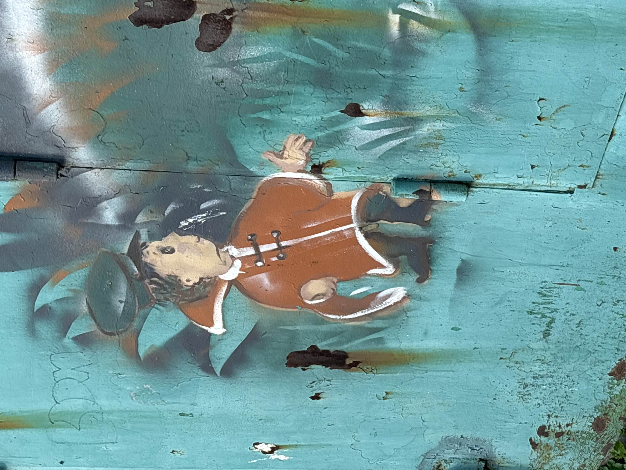 The image shows a close-up, eye-level shot of graffiti on a weathered, teal-colored metal surface, possibly a door or gate. The graffiti depicts a cartoon-like figure in historical attire, with an orange tunic, white trim, and a dark hat. The figure's right arm is extended outwards in a gesture. The background of the graffiti features abstract spray-painted elements in shades of green, brown, and black, with a distinctive shadow effect.
The metal surface exhibits significant signs of age and wear, including rust spots and peeling paint, particularly noticeable around the graffiti and along the vertical seam of what appears to be a hinged door. The color palette of the graffiti is vibrant against the muted tones of the decaying metal.
While the image is a close-up and lacks contextual environmental cues, the presence of graffiti, rust, and peeling paint suggests an urban setting. Based on the provided information, this graffiti is located in Novosibirsk, Russia. The overall impression is one of urban decay and artistic expression.