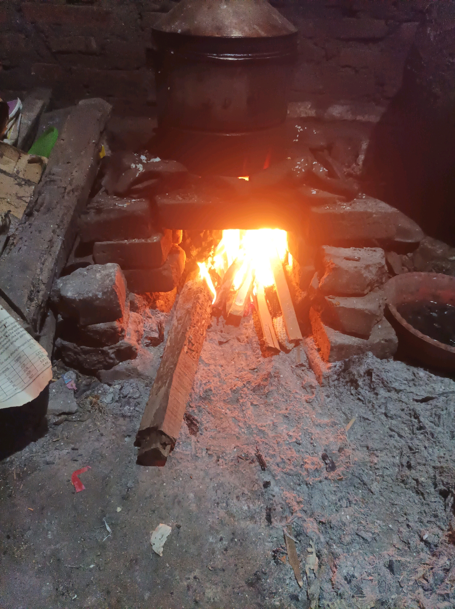 This image depicts a traditional outdoor kitchen in Canga'an, Indonesia, centered around a brick stove with a fire burning within its hearth. Several pieces of firewood are arranged in the fire, with flames clearly visible. Above the hearth, a large metal pot rests on the brick structure, suggesting food preparation is in progress. The stove itself is constructed from rough, uneven bricks and stone, with ashes and embers spread around the base, indicating recent use. To the left of the stove, a stack of rough-hewn wooden beams is visible, along with a crumpled piece of paper that appears to be a document or newspaper. The ground is composed of packed dirt and ash, with some scattered debris including small pieces of red and white paper. To the right of the stove, a dark-colored ceramic bowl, possibly containing liquid, is present. The overall environment appears rustic and utilitarian, consistent with a traditional cooking setup.