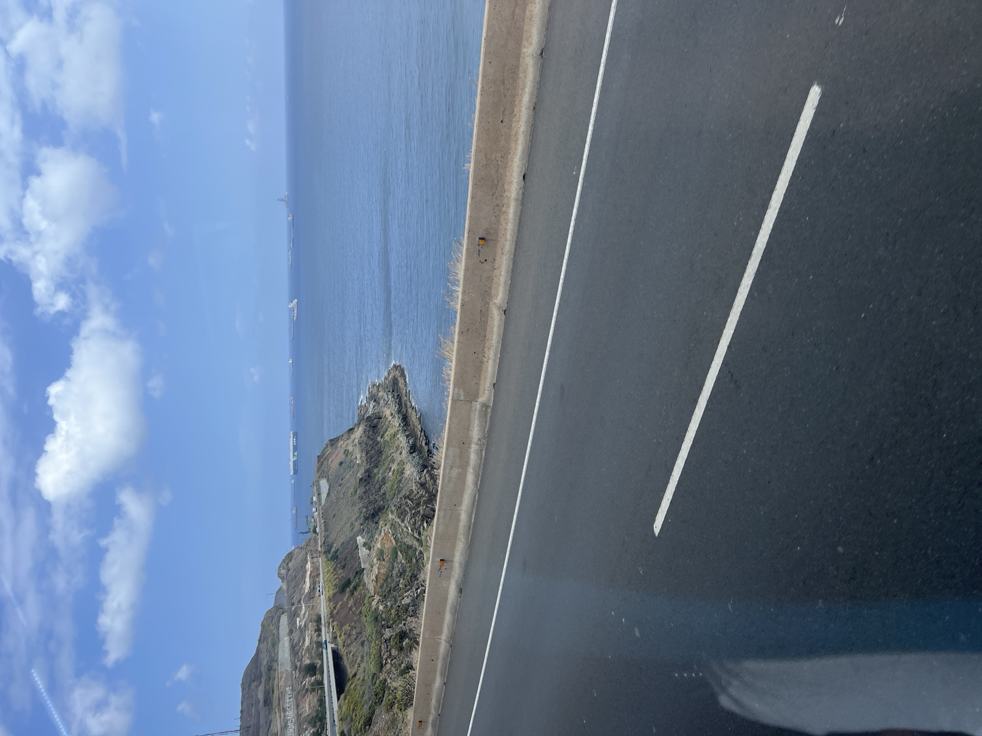 The image captures a coastal landscape under a bright, clear sky, likely from a vantage point on a road. The scene is dominated by a dark asphalt road in the foreground, featuring a solid white line on its inner edge and a broken white line further towards the center. A concrete barrier separates the road from a steep, rocky cliff face that descends to the ocean below.
Several small, yellow reflective markers are embedded along the top of the concrete barrier, with some dry grass and weeds growing from its base. The cliff face is rugged, covered sparsely with vegetation, and shows signs of human alteration or infrastructure further down. Notably, white text is painted onto the lower part of the cliff, reading "NO HAY PLANETA B" (Spanish for "There is no planet B"), suggesting an environmental message.
Beyond the cliff, the vast, blue ocean stretches to the horizon. Several large ships, some appearing to be cargo or container vessels (one with green markings is visible), are scattered across the distant waters, suggesting a busy shipping lane or port area. The sky above is a clear blue with scattered white cumulus clouds, indicating a sunny day. The overall impression is one of a scenic coastal drive or overlook, characteristic of a location like Las Palmas, Spain, given its prominent port and rugged coastline. The time of day appears to be midday or early afternoon due to the bright, direct sunlight.