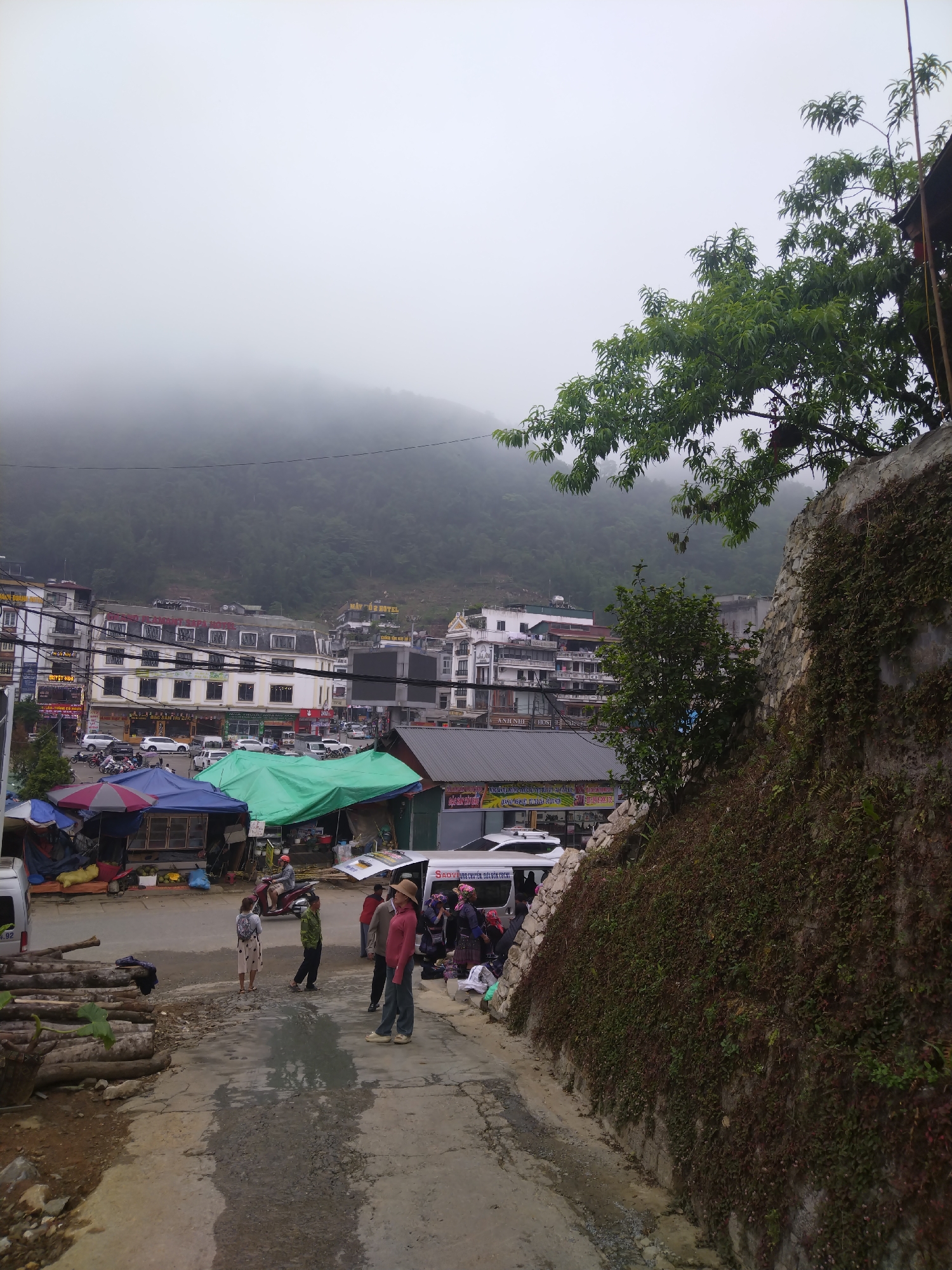 The image depicts a street scene in a mountainous area, potentially a town in a fog-shrouded region. In the foreground, a paved road slopes down into the scene, with a stone and vegetation-covered wall on the right and a stack of logs on the left. Several people are gathered in the middle distance, some standing and conversing, others appearing to be vendors with goods displayed. Market stalls and vehicles are visible in the background behind the group of people. Buildings, predominantly multi-story structures, line the upper part of the image, and a densely forested mountain fades into the fog in the distance. The weather appears overcast and damp.