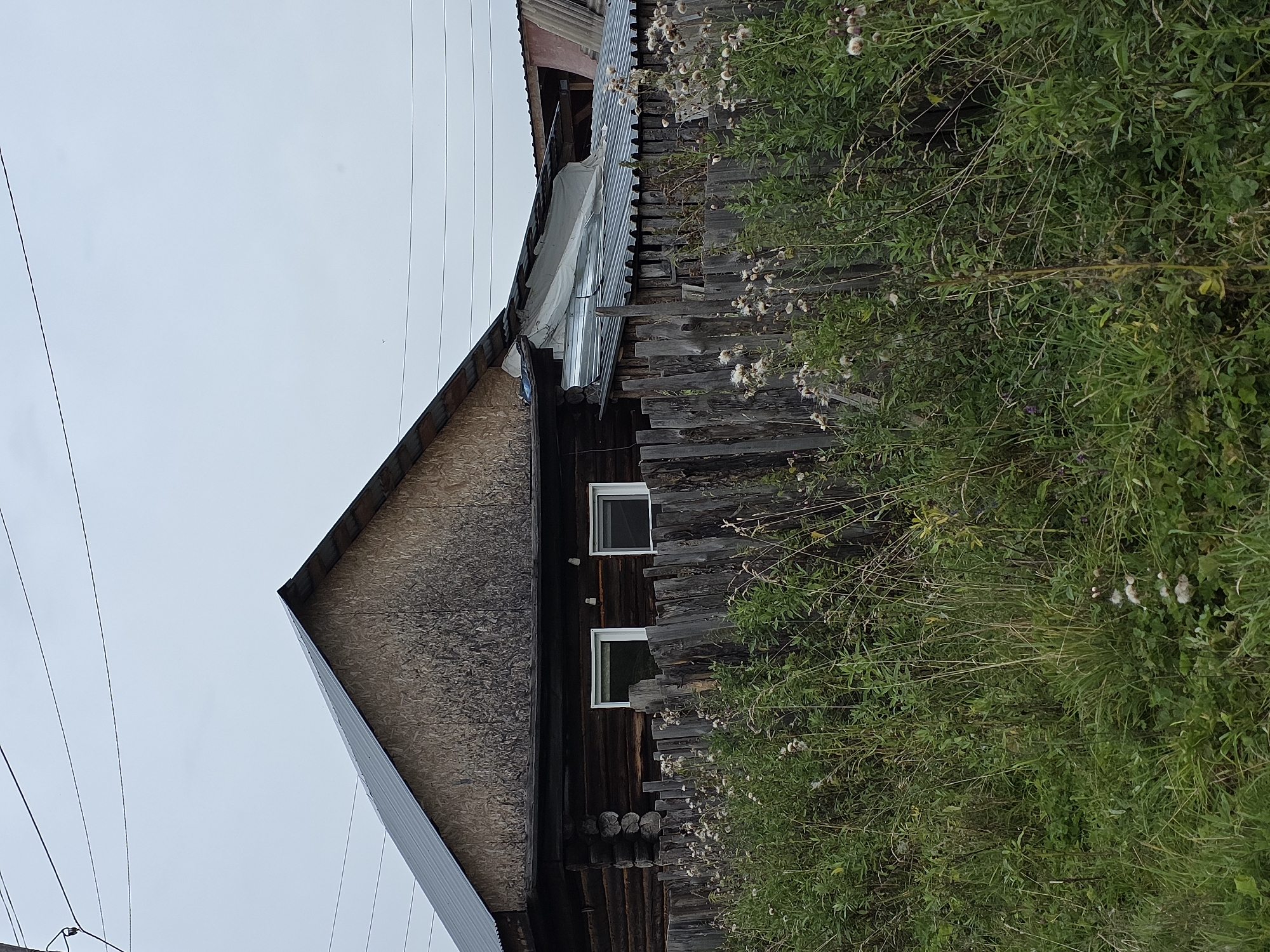 The image depicts a weathered, wooden log house with a metal roof, situated behind a rustic wooden fence. The house has two windows with white frames visible on the front wall. The gable end of the house is covered with a rough, textured material that appears to be wood chipboard, and it shows signs of staining or weathering. Adjacent to the main house, a section of the roof appears to be made of corrugated metal, with some areas covered by a white tarp. In front of the fence, tall green grass and weeds, some with fluffy seed heads, grow densely. Overhead, power lines stretch across the frame against a pale, overcast sky. The scene suggests a rural or semi-rural setting. The mention of Magnitka, Russia, places this scene within a specific geographic context, likely a residential area within or near the city.