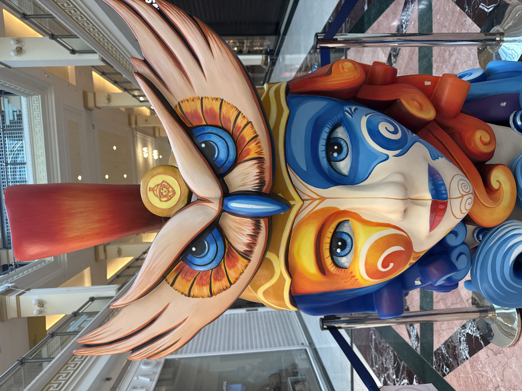 This is a photograph of a large, colorful sculpture of a face with owl-like features, located in an indoor marketplace or mall. The sculpture is centrally framed and dominates the image. It features a prominent owl headpiece with large, stylized wings extending upwards and outwards. The owl's eyes are a vibrant blue with orange and black accents, and its beak is a striking blue. Below the owl head, the sculpture depicts a human-like face divided down the middle with contrasting colors. The left side of the face is painted in shades of orange, yellow, and white with swirling patterns, while the right side is a deep blue with similar white swirl designs. The face has bright blue eyes, red lips, and red and blue hair or headdress elements that curl outwards. The sculpture is positioned in front of a blurred background of architectural features common to indoor shopping environments, including columns, railings, storefronts, and lighting. Blue stanchions with dark blue velvet ropes are visible in the foreground, indicating a roped-off area around the sculpture. The floor is a patterned marble.