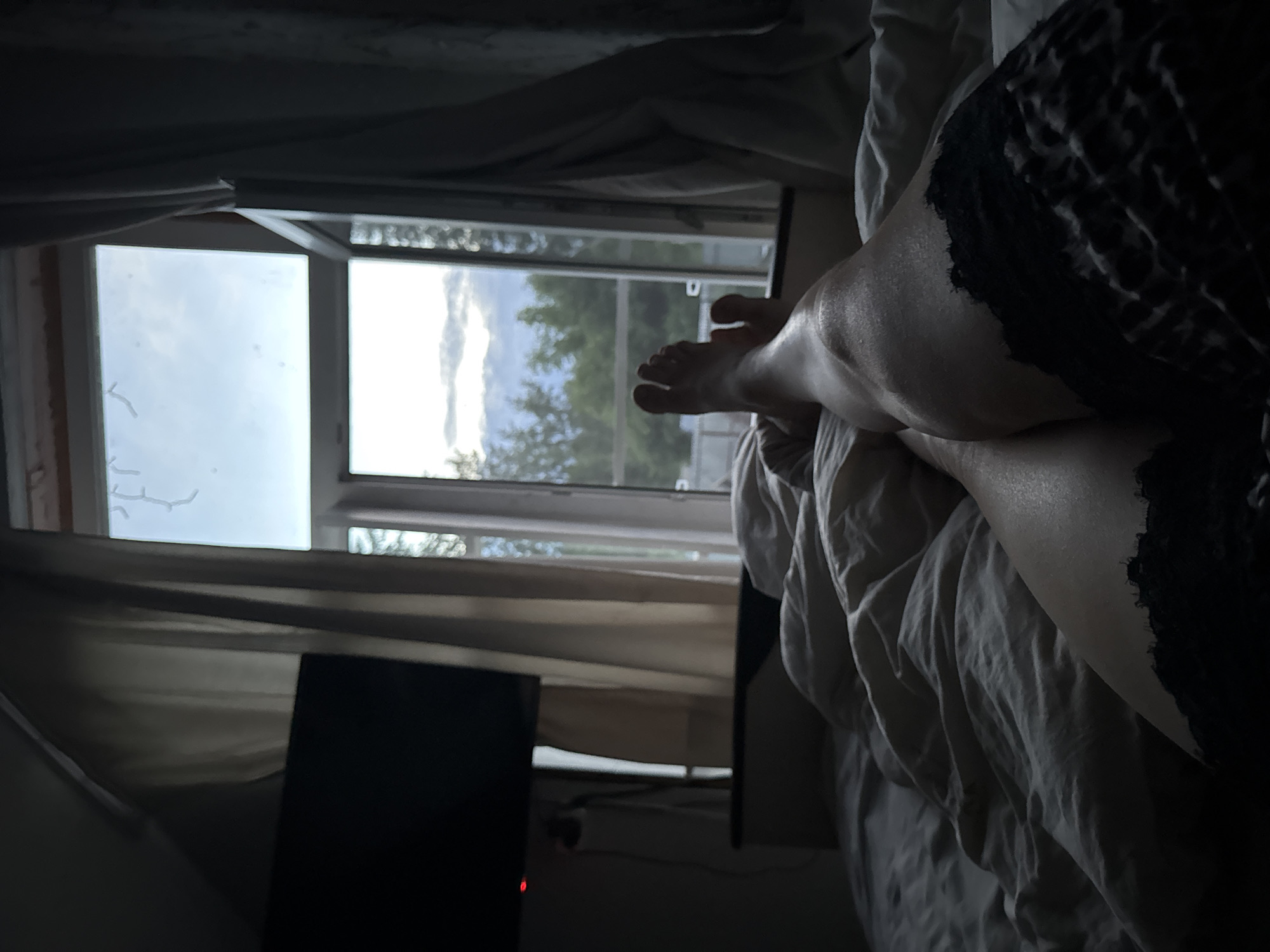 A person's legs are visible in the foreground, resting on a bed with rumpled white sheets. The person is wearing black lace underwear or stockings. To the left, a black television is mounted on the wall above a dark surface. Behind the bed, a window is open, revealing a view of trees and a cloudy sky. Light-colored curtains hang on either side of the window. The scene appears to be indoors, likely in a bedroom, and based on the geographical information provided, it is located in the city of Chaikovsky, Russia.