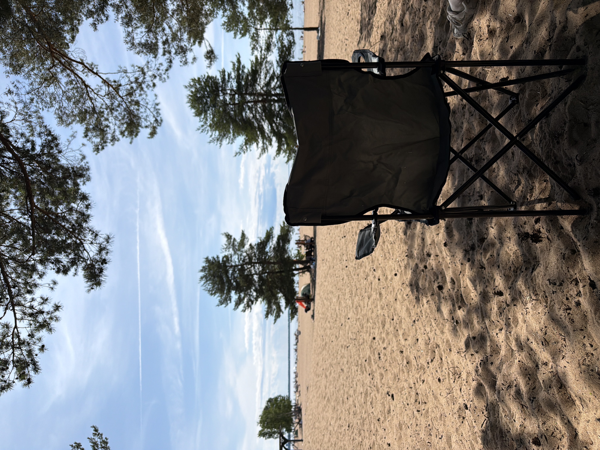 This media file depicts a beach scene on a sunny day, likely in the afternoon, given the shadows. The foreground is dominated by a dark-colored camping chair, casting a shadow on the sandy ground. The sand has some debris and appears slightly rippled. In the background, tall pine trees are visible, characteristic of a coastal or lakeside environment. Beyond the trees, a body of water, likely a lake or the sea, can be seen with a distant shoreline and several people scattered along the beach, some appearing to be sitting or standing. A few tents are also visible. The sky is blue with scattered white clouds, and a contrail from an aircraft is faintly visible. The image is taken from a low angle, looking upwards towards the sky and the trees. The overall mood is peaceful and relaxed, suggesting a leisurely day spent outdoors. There is no visible text in the media file. The location context of Solnechnoye, Russia, aligns with the presence of pine trees and a beach environment.