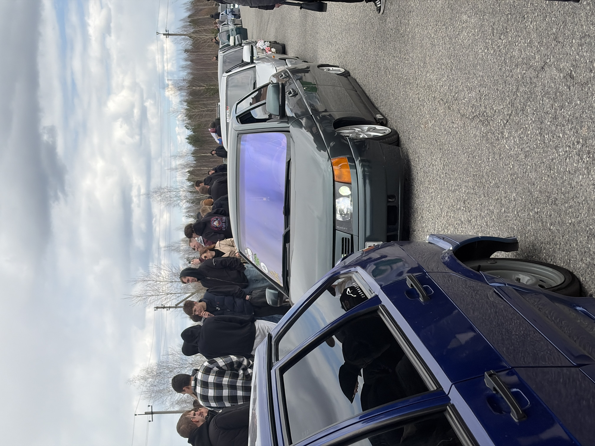 The image shows a gathering of people and cars on an asphalt surface under a cloudy sky. The cars appear to be parked in a row, with a dark blue car in the foreground and several other cars lined up behind it, varying in color and model. Some people are standing next to or near the cars, seemingly interacting with one another. The background includes trees and power lines, indicating a location perhaps on the outskirts or a less densely populated area. The license plate on the windshield of the gray car indicates a car registered in Perm, Russia.