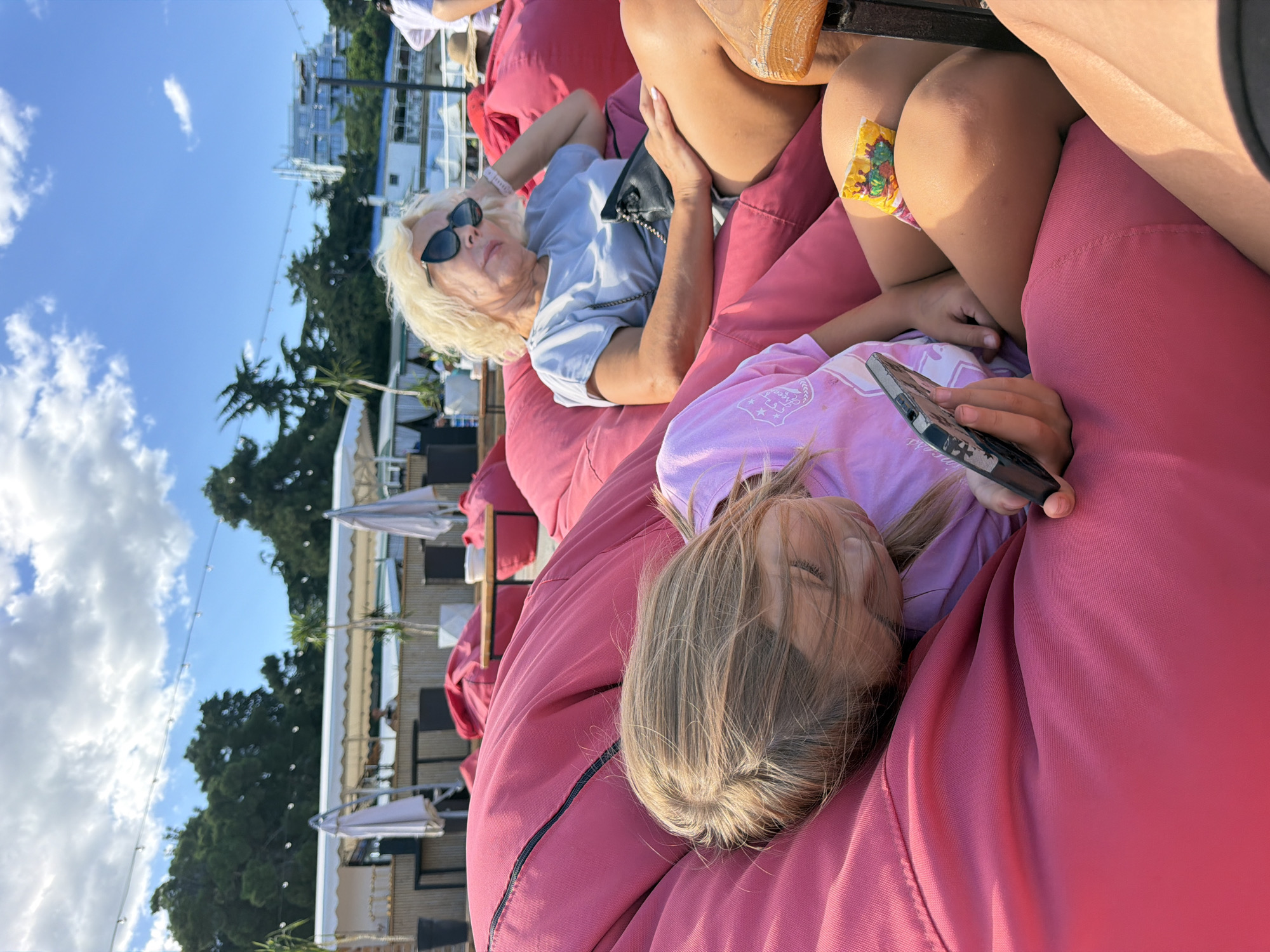 In Alushta, Ukraine, on a sunny day, two individuals are relaxing on oversized pink beanbag chairs at an outdoor venue. In the foreground, a young girl with blonde hair is lying down and looking at her mobile phone. She is wearing a pink t-shirt. Next to her, an older woman with blonde hair, wearing sunglasses and a light blue t-shirt, is resting with her arm behind her head. She has a black purse on her lap. In the background, there are more beanbag chairs, tables, umbrellas, and trees, suggesting a beachside or outdoor cafe setting. The sky is blue with scattered clouds, and some buildings are visible in the distance.