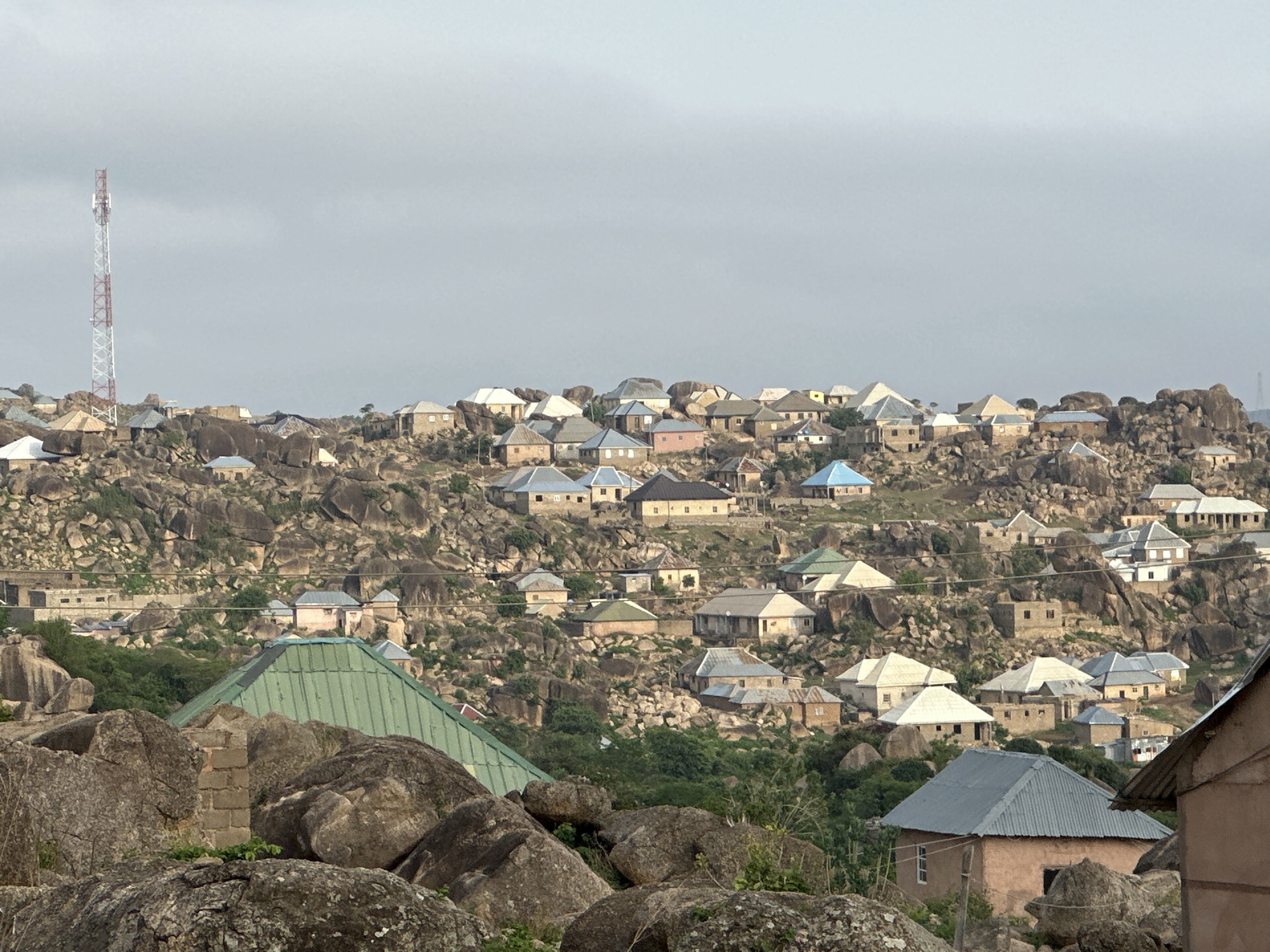 The image depicts a densely populated residential area situated on a rocky hillside in Jos, Nigeria. The houses are primarily single-story structures with corrugated metal roofs in various colors including silver, green, and blue. The architecture appears simple and functional, with the houses nestled among large granite boulders that dominate the landscape. A communications tower is visible in the upper left of the frame. The sky is overcast, and the overall lighting is diffused. The foreground features large boulders and sparse vegetation, suggesting a rugged terrain.