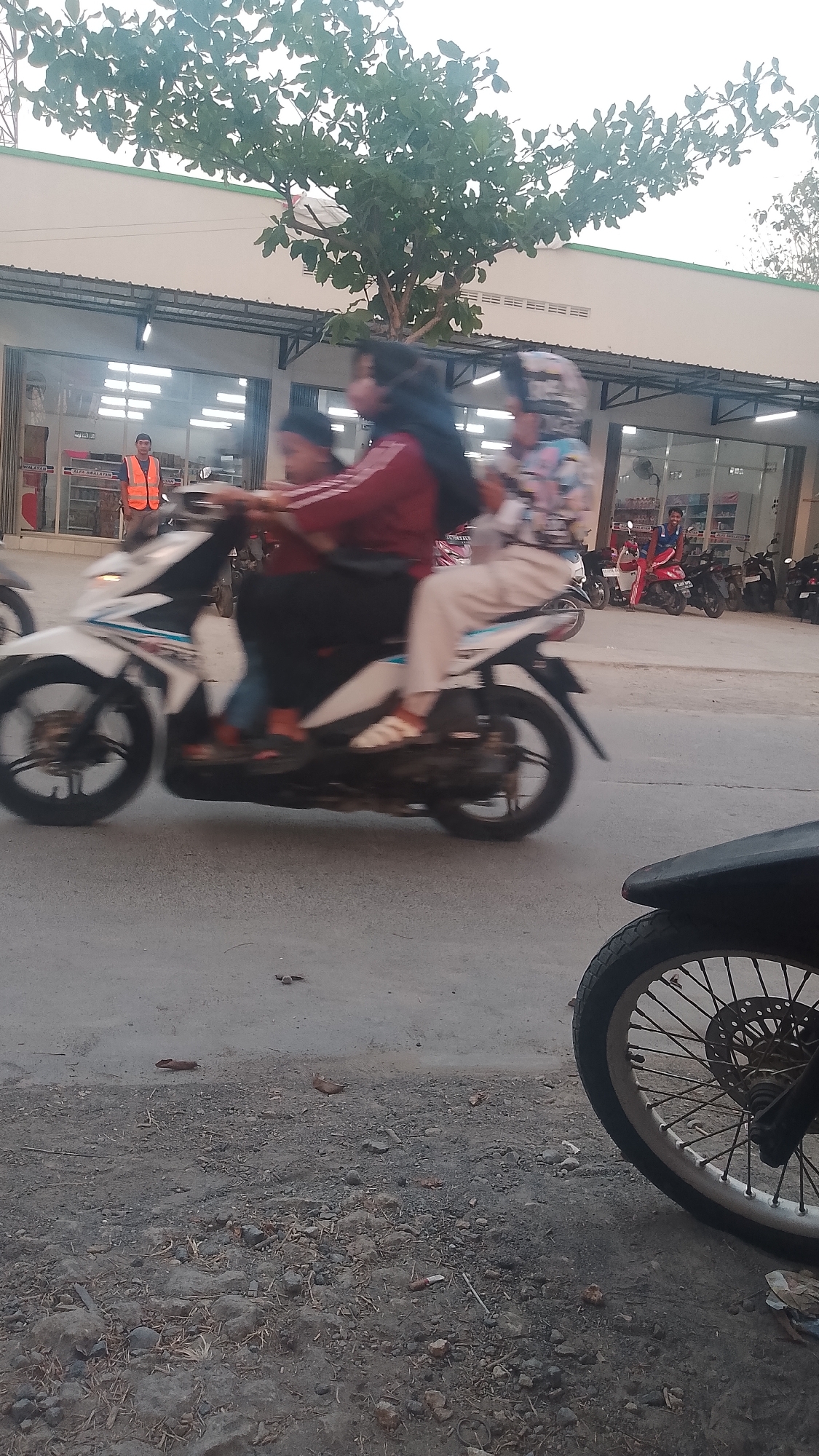 A medium shot captures a blurred scene of a white motorcycle carrying three people traveling on a paved road in front of a row of shops. The driver, a person wearing a red jacket and dark pants, is at the front. A person in a patterned hijab and light-colored pants is seated behind the driver, and a third person, also in a patterned hijab and light-colored pants, is at the very back. The shops in the background have open glass fronts, revealing shelves stocked with products. To the left of the motorcycle, a person in an orange vest stands near the entrance of one of the shops. The environment appears to be an urban setting, likely in the city of Karanglangu, Indonesia, given the presence of motorcycles and a general street scene common in Indonesian cities. The foreground shows a rough, gravelly shoulder with some debris. The lighting suggests it is daytime, possibly late afternoon.