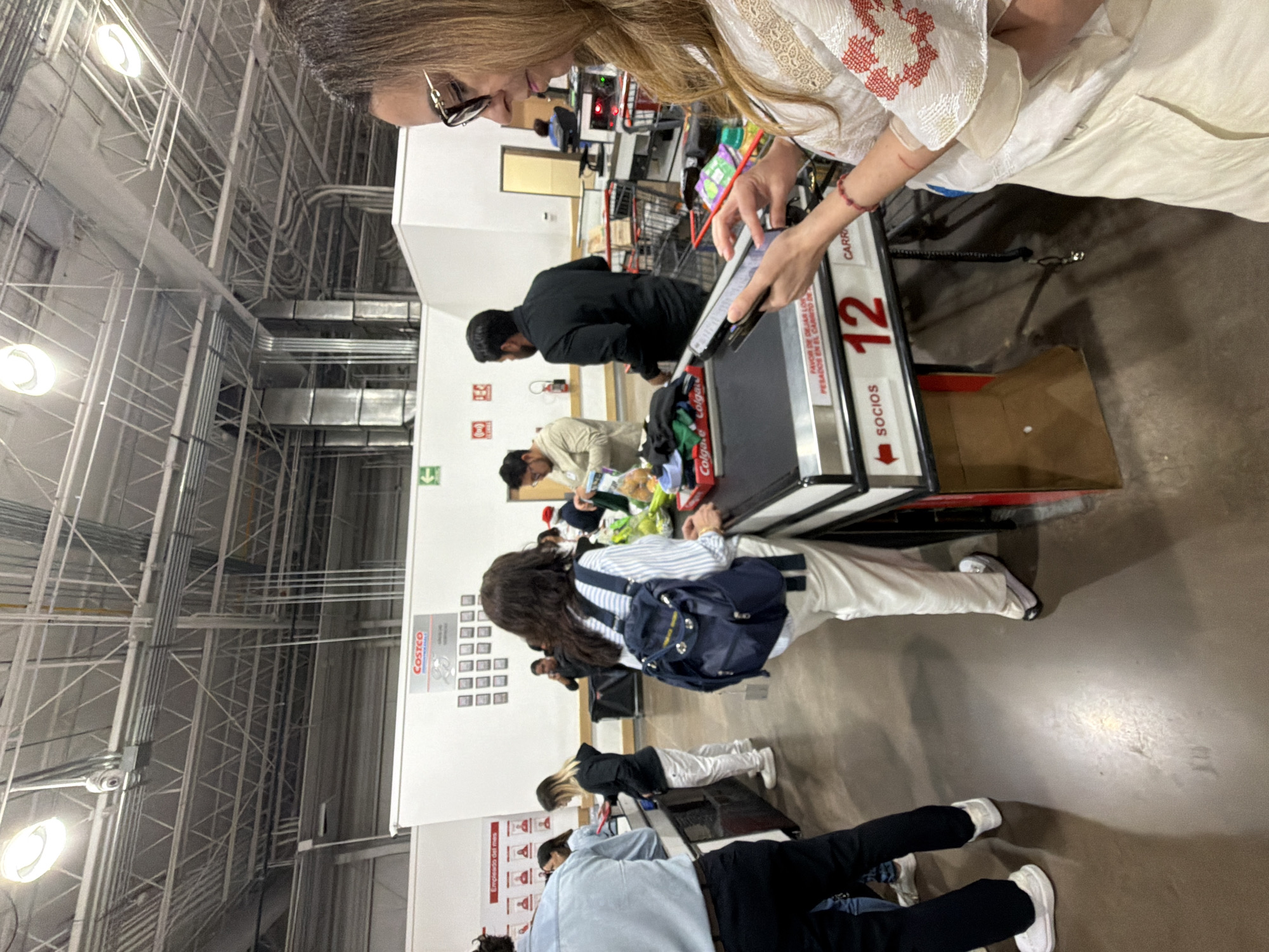 A scene at Costco in Puebla, Mexico, showing several people at checkout lanes. The foreground features a woman with long brown hair, wearing glasses and a white embroidered blouse, looking at her phone while standing at checkout lane 12. A customer in a black shirt is unloading items onto the conveyor belt. Further down the lane, another customer in a light-colored shirt is also at the checkout. A woman with a blue backpack is visible in the adjacent checkout lane. In the background, there are additional people waiting in line at other checkout lanes. The environment shows the typical Costco warehouse setting, with high ceilings and industrial lighting. A "Costco" sign is visible on a nearby wall, along with a "Empleado del mes" (Employee of the Month) display. A red sign with "SOCIOS" and an arrow pointing to the left is visible, suggesting the direction of the checkout line. The floor is concrete.
