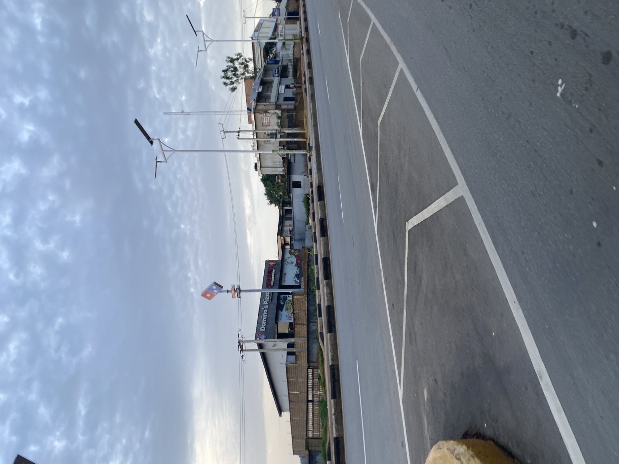 The image depicts a street scene in Jos, Nigeria, under a cloudy sky. A highway with a distinct yellow and black curb runs horizontally across the foreground. White lines mark the lanes and a parking area on the right side of the road. In the background, a Domino's Pizza and Cold Stone Creamery establishment is visible, identifiable by its signage. Several streetlights, some with solar panels, line the roadside. The buildings in the background appear to be a mix of commercial and residential structures, some showing signs of wear. The overall environment suggests an urban setting, possibly during the early morning or late afternoon due to the diffused light. No people are clearly visible, and no specific actions or interactions are captured.