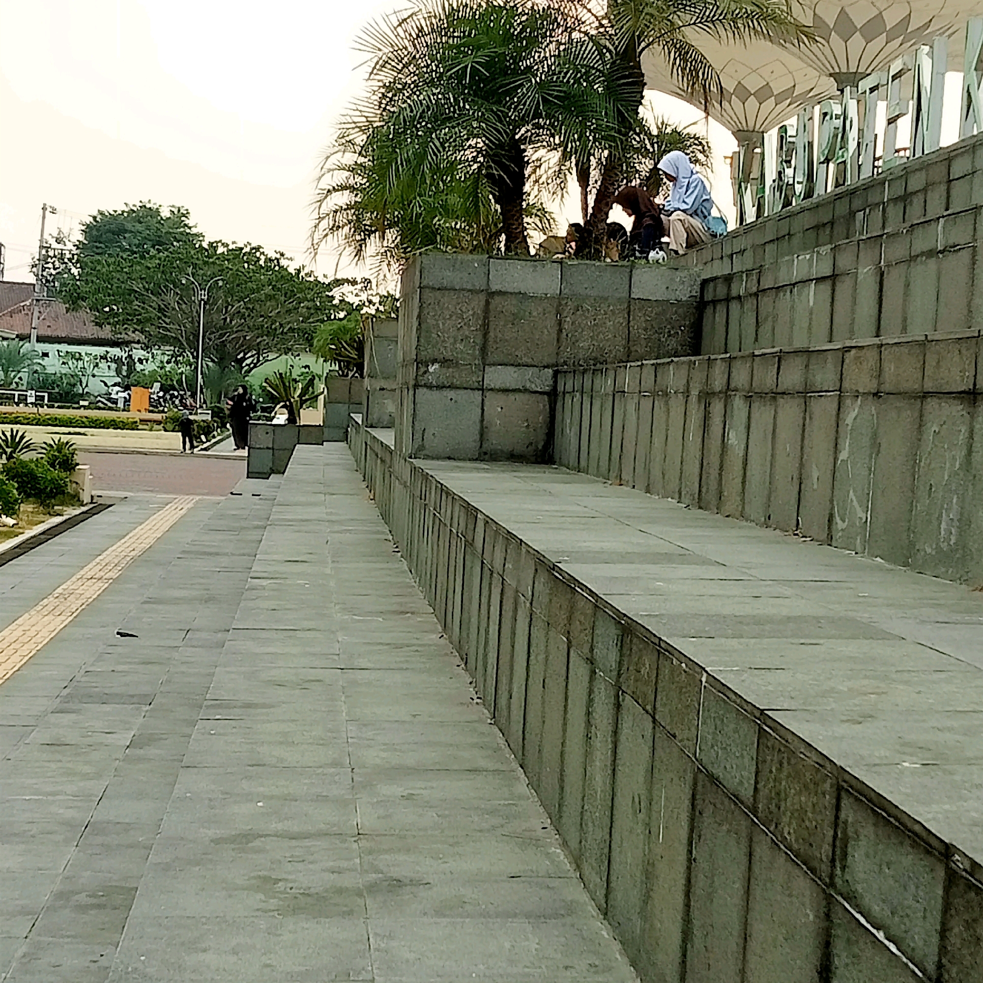 The image depicts an outdoor scene in Karanganyar, Indonesia, likely near a modern building with distinctive white and grey patterned canopies overhead. In the foreground, a wide pathway paved with grey tiles leads diagonally into the frame from the left. A tactile paving strip, yellow and grey, runs along the left edge of this pathway. To the right of the pathway, a raised, tiered structure made of large, grey stone blocks is visible. Several people are seated on the top tier of this structure. One person wears a light blue hijab and tan pants, and appears to be looking at a mobile device. Next to them, another individual in darker clothing is also seated, and there are a couple of small animals, possibly dogs, nearby. In the middle ground, on a lower level, two figures are walking on a paved area. One is dressed in dark clothing and appears to be standing, while the other, in dark clothing, is walking away from the camera. Behind them, there is greenery, including trees and shrubs, and a building with a red roof. The sky above is overcast and pale. The overall impression is a public outdoor space, possibly a plaza or an area adjacent to a commercial or civic building.