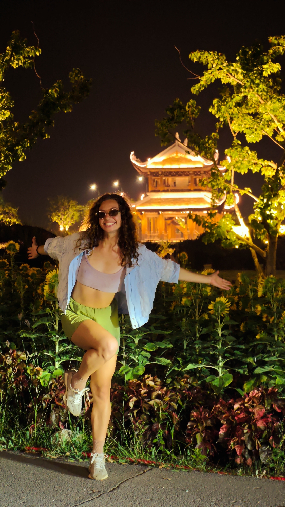 A full-body shot captures a woman posing with her arms outstretched in front of a brightly lit pagoda at night. The woman stands on a paved path with a bed of sunflowers and other foliage to her right. She has curly brown hair, sunglasses, and is wearing a light purple sports bra, green shorts, and a light blue oversized shirt. Her left leg is bent and crossed over her right, and she is giving a thumbs-up with her left hand. The pagoda, with traditional East Asian architecture, is illuminated by warm, yellow lights, and several trees with glowing leaves frame the scene. The background is dark, indicating it is nighttime.