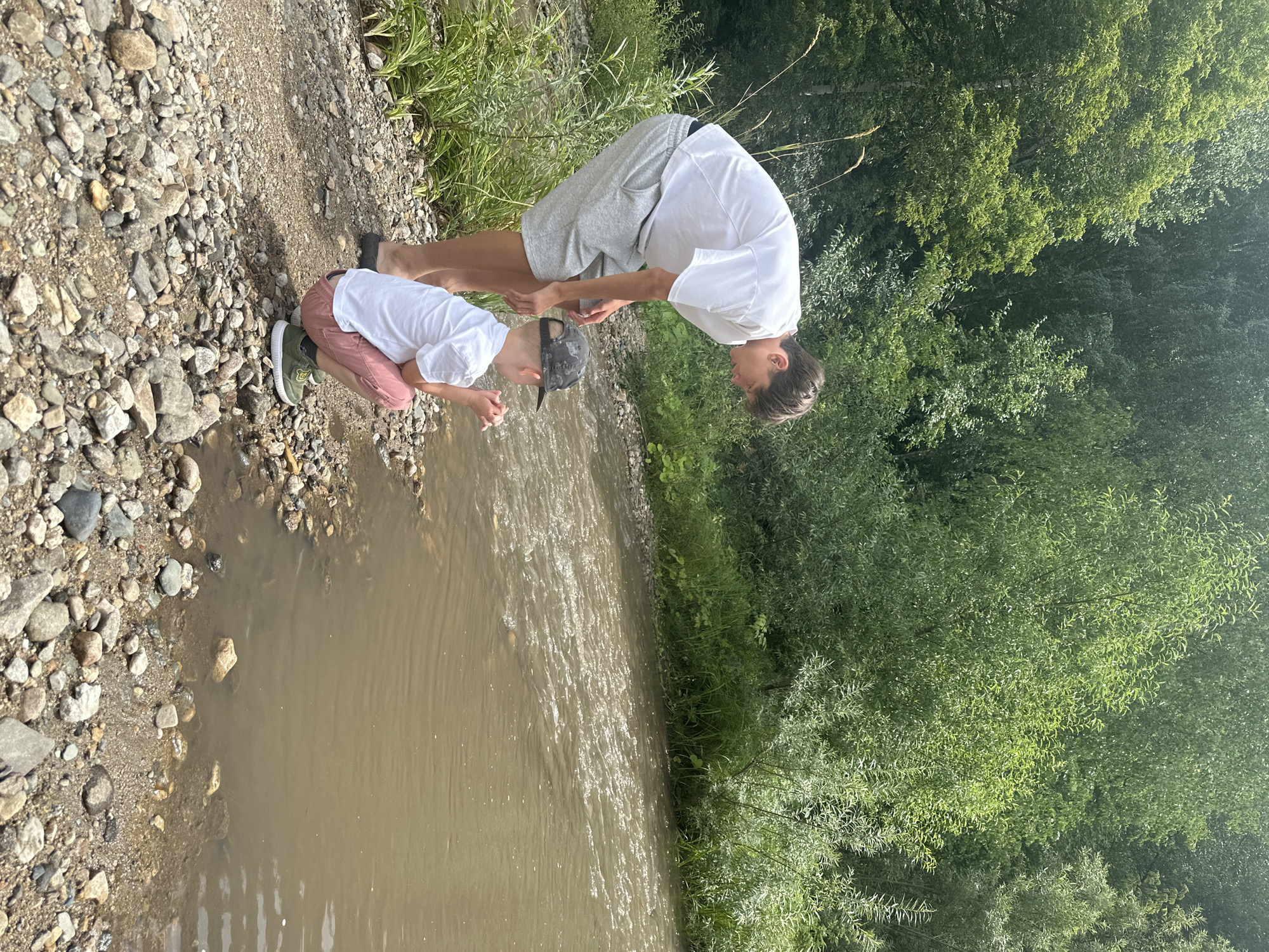 Two individuals, likely brothers, are by a shallow, muddy river. The taller individual, possibly a teenager, is wearing a white t-shirt and grey shorts and is bending over to observe the water. The younger individual, wearing a white t-shirt, pink shorts, a baseball cap, and green shoes, is squatting near the water's edge, appearing to play with their hands in the water. The riverbank is covered in small stones and pebbles. Lush green trees and foliage surround the area, suggesting a natural, rural setting. The location is identified as Danilovka (Altai), Russia.