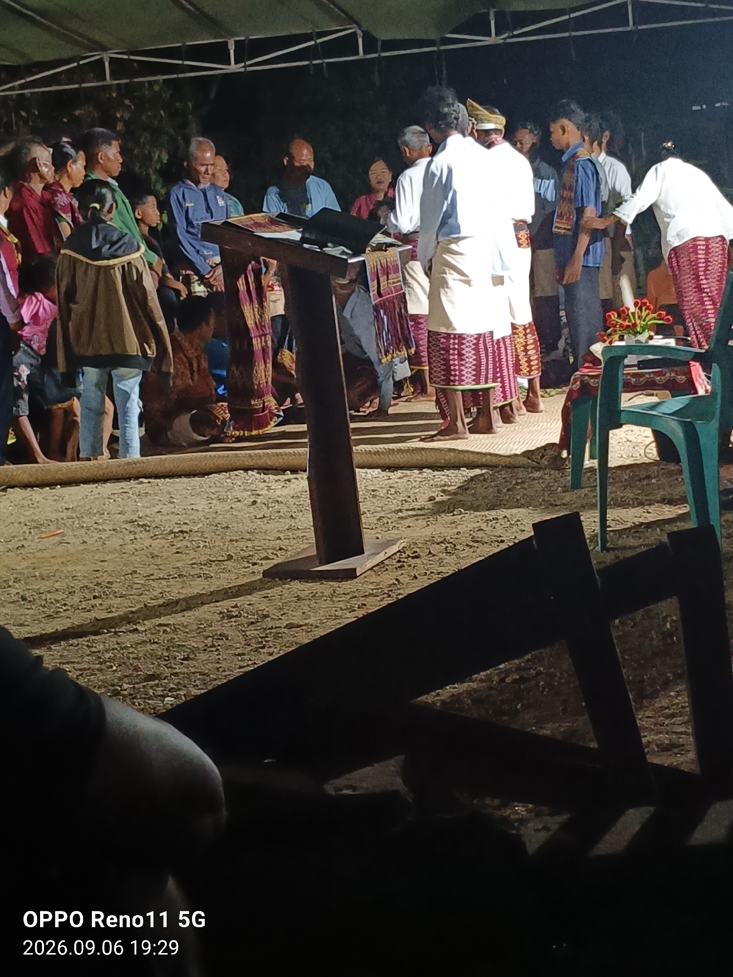 In Otan, Indonesia, an outdoor gathering is taking place under a canopy. A group of people, dressed in traditional attire, are standing around a wooden lectern. Some are listening attentively, while others appear to be participating in a ceremony. The ground is covered with gravel, and in the foreground, two dark, wooden structures cast long shadows. A green plastic chair with a floral arrangement sits to the right of the lectern. The lighting suggests it is evening.