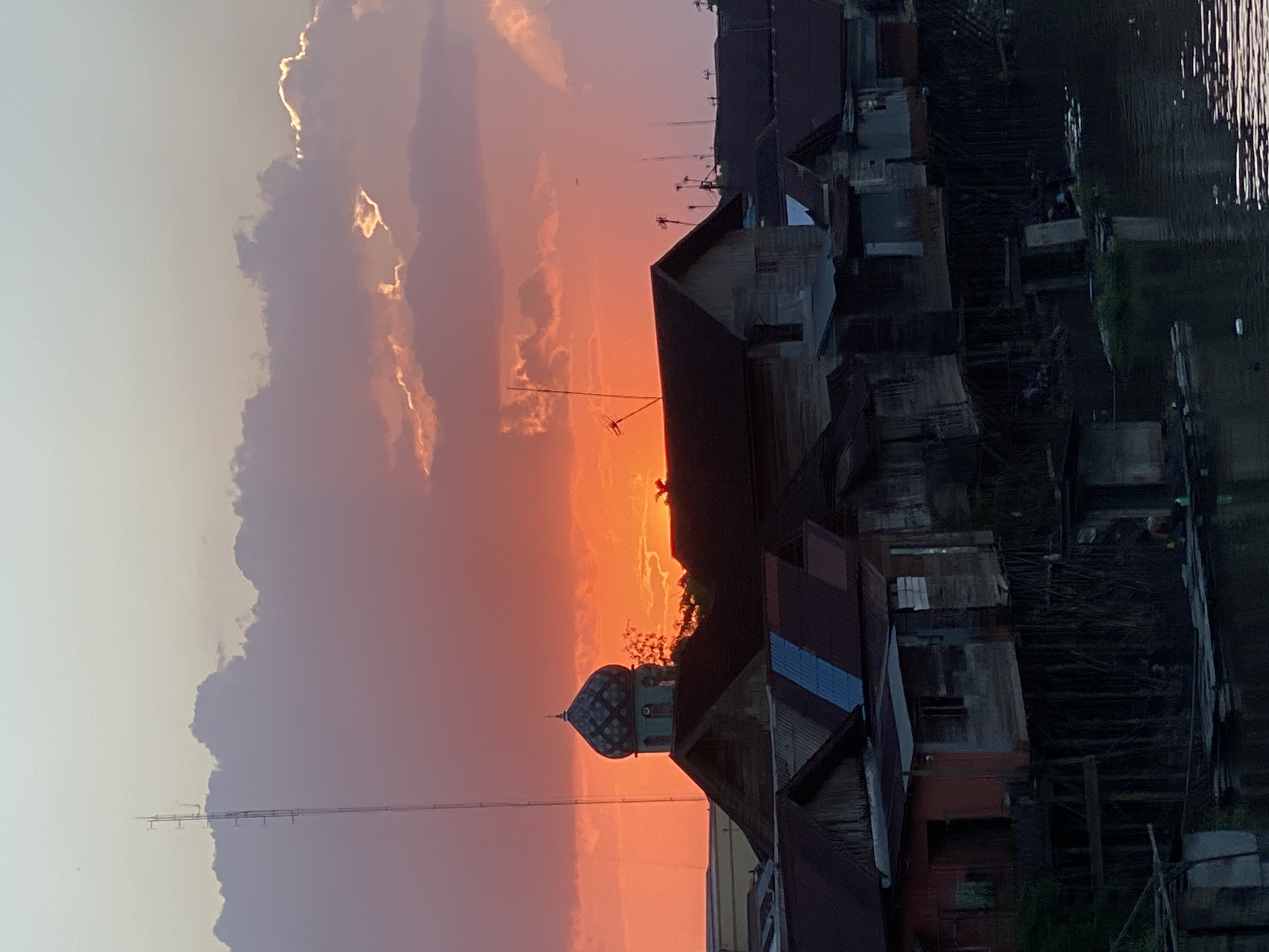 The image captures a sunset over Tunggul Irang Ulu, Indonesia. The sky is ablaze with vibrant orange and pink hues, with dramatic clouds framing the scene. In the foreground, a cluster of stilted houses lines a body of water. The architecture is simple and weathered, with corrugated metal roofs and wooden walls. A prominent dome-topped building, possibly a mosque, stands out amidst the houses. A tall communication tower is visible in the background, adding a modern element to the otherwise traditional setting. The water reflects the warm colors of the sky, creating a serene and picturesque atmosphere.